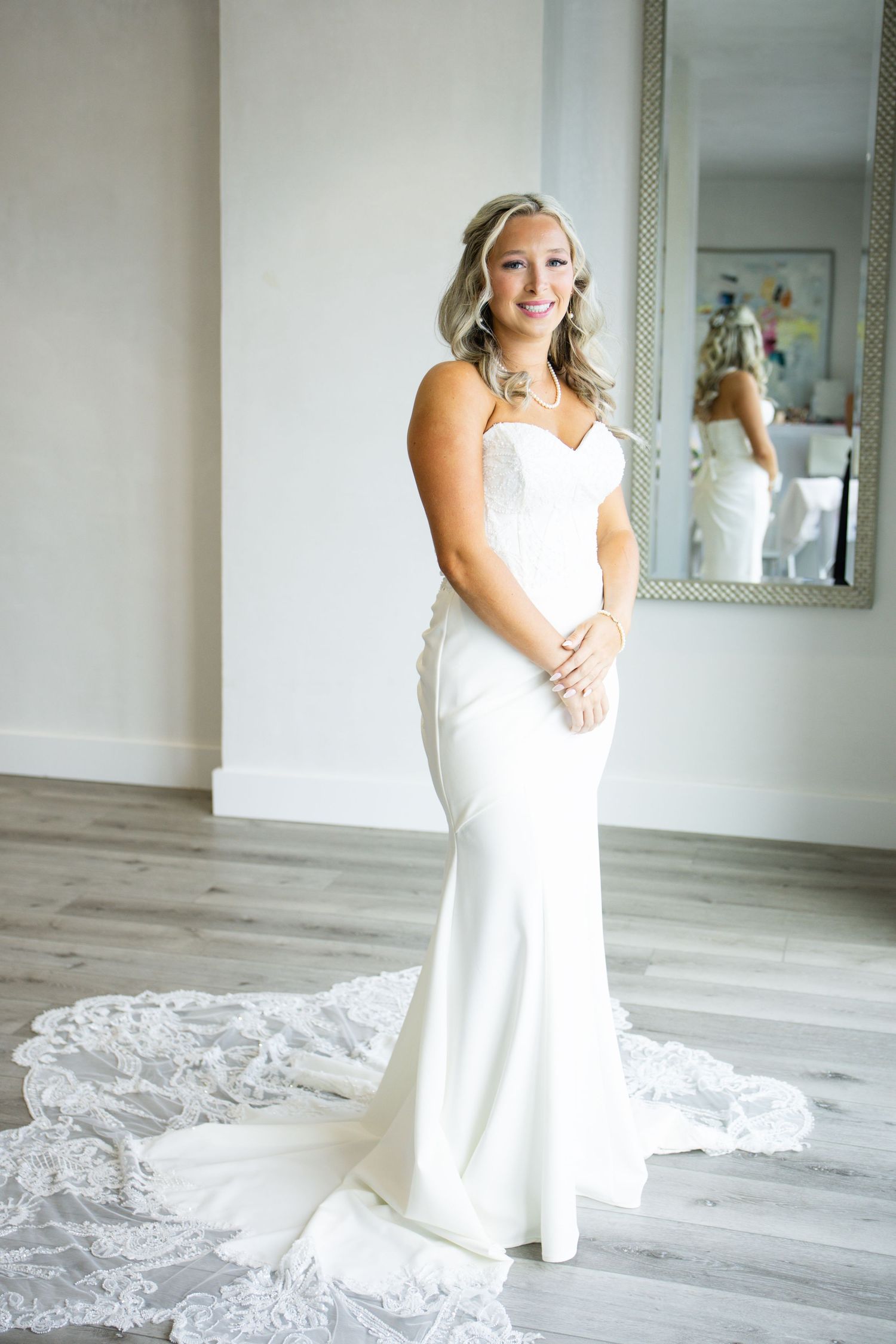 Bride in elegant strapless white mermaid gown with lace train smiles in bridal boutique with mirror reflection.