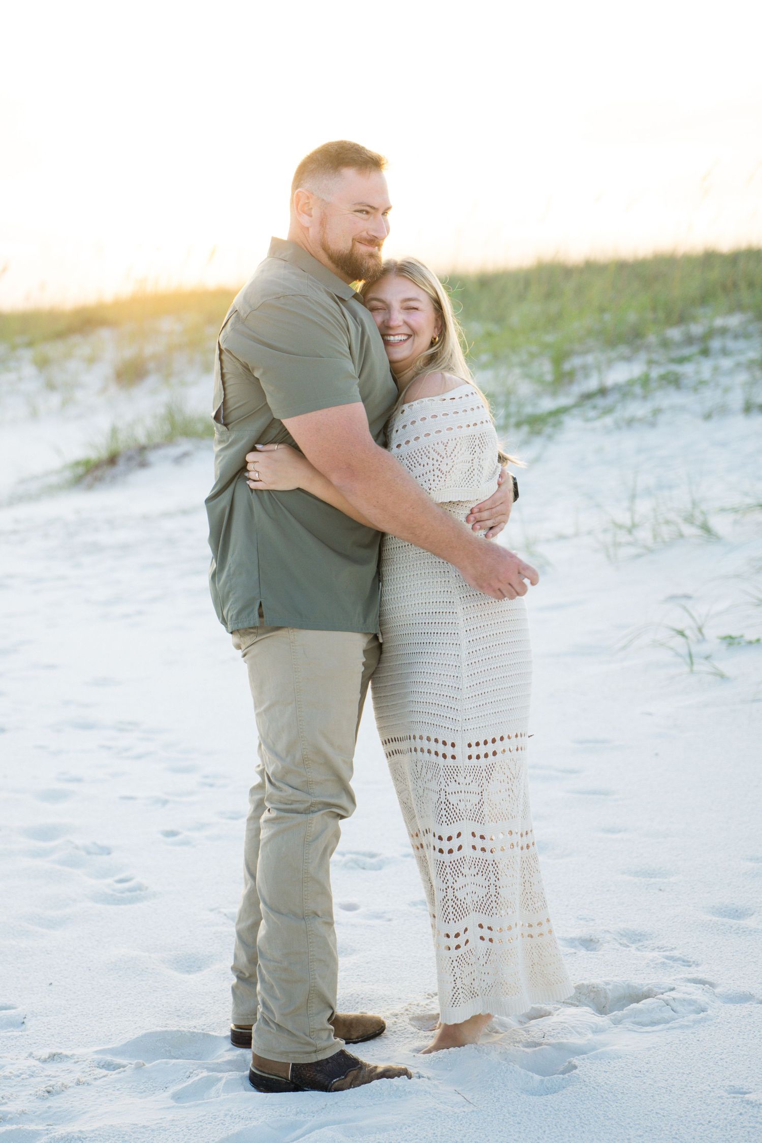 Couple embracing on sandy beach at sunset, woman in white crochet dress, man in green shirt and khakis.