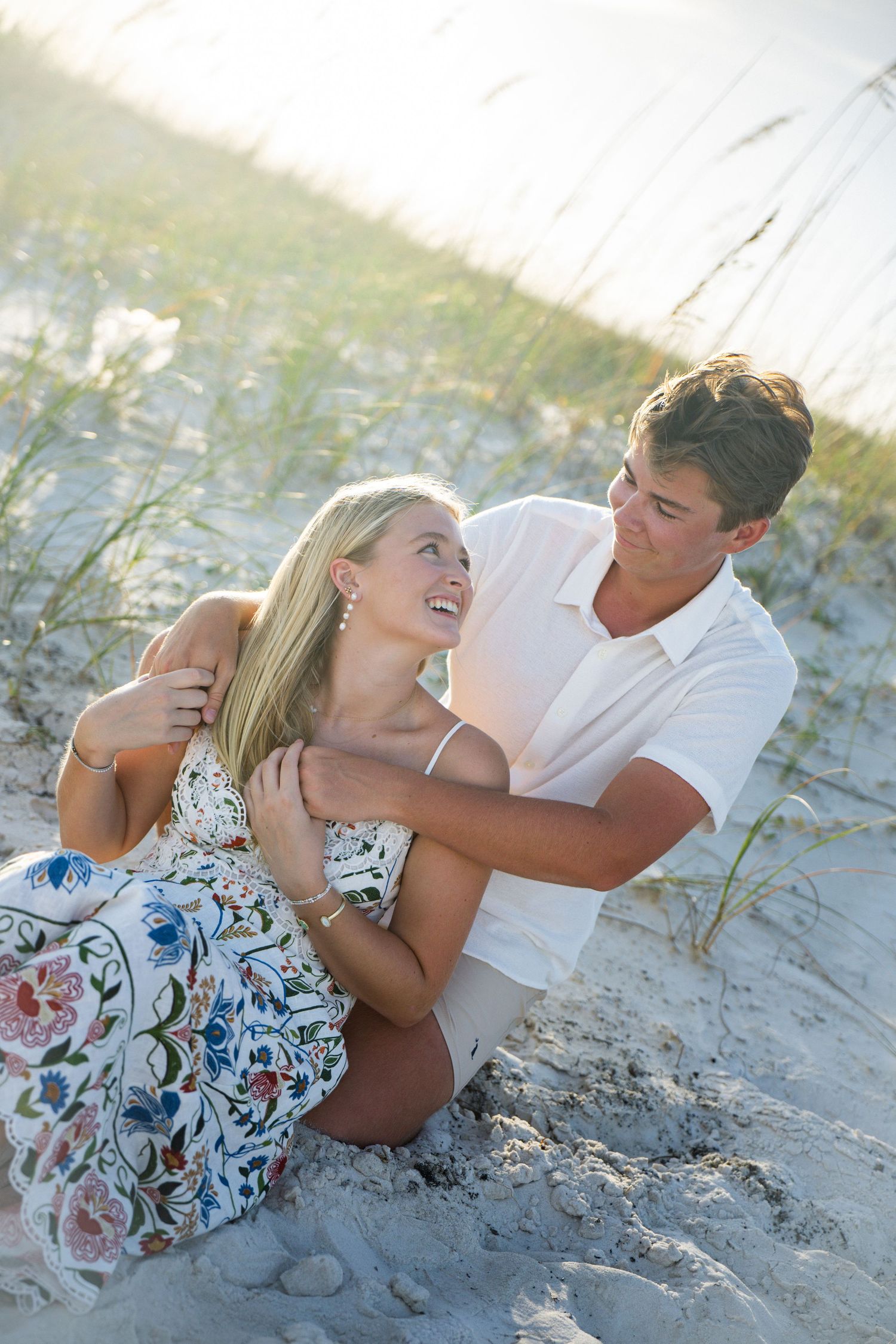 A happy young couple sits together on a sandy beach, sharing a romantic moment near sea oats in warm sunlight.
