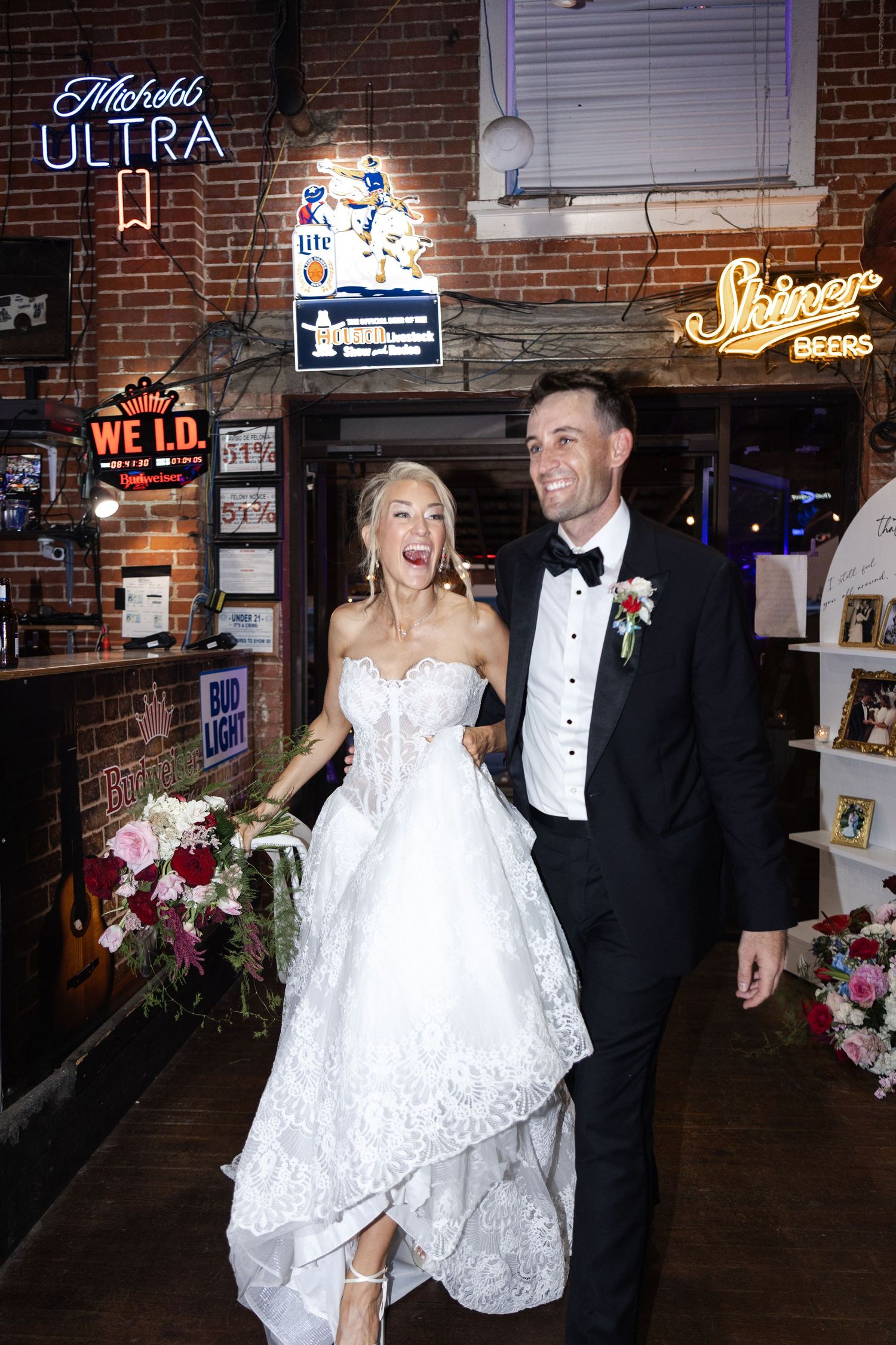 Joyful bride in lace gown and groom in tuxedo making grand entrance at wedding reception venue with neon beer signs.