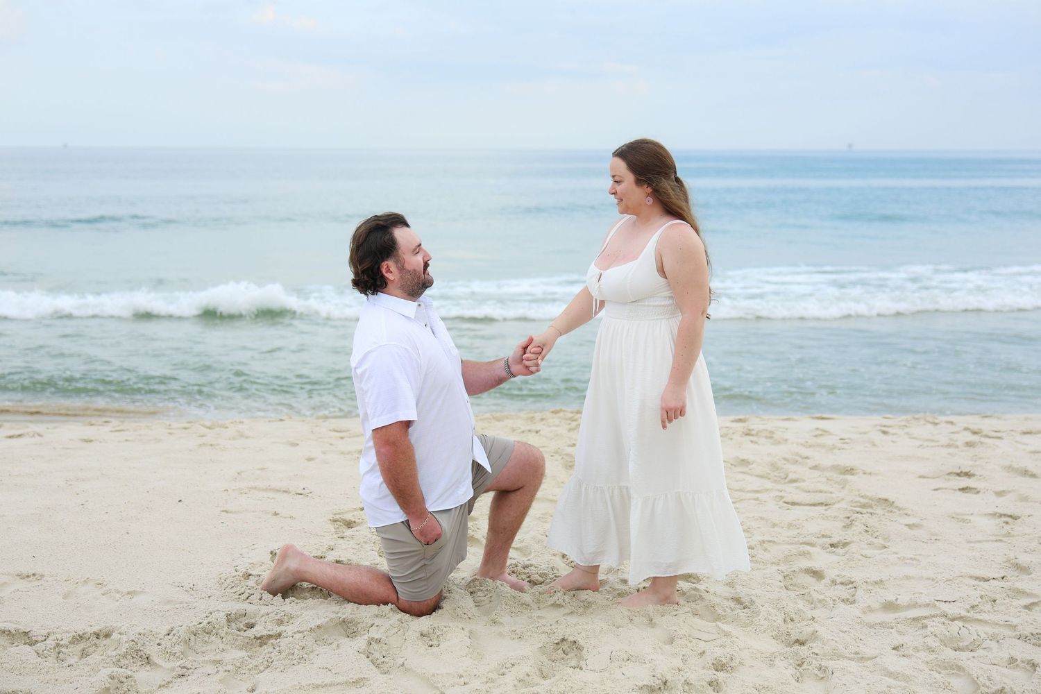 Man proposes to woman on one knee on a sandy beach, holding hands with ocean waves in the background.