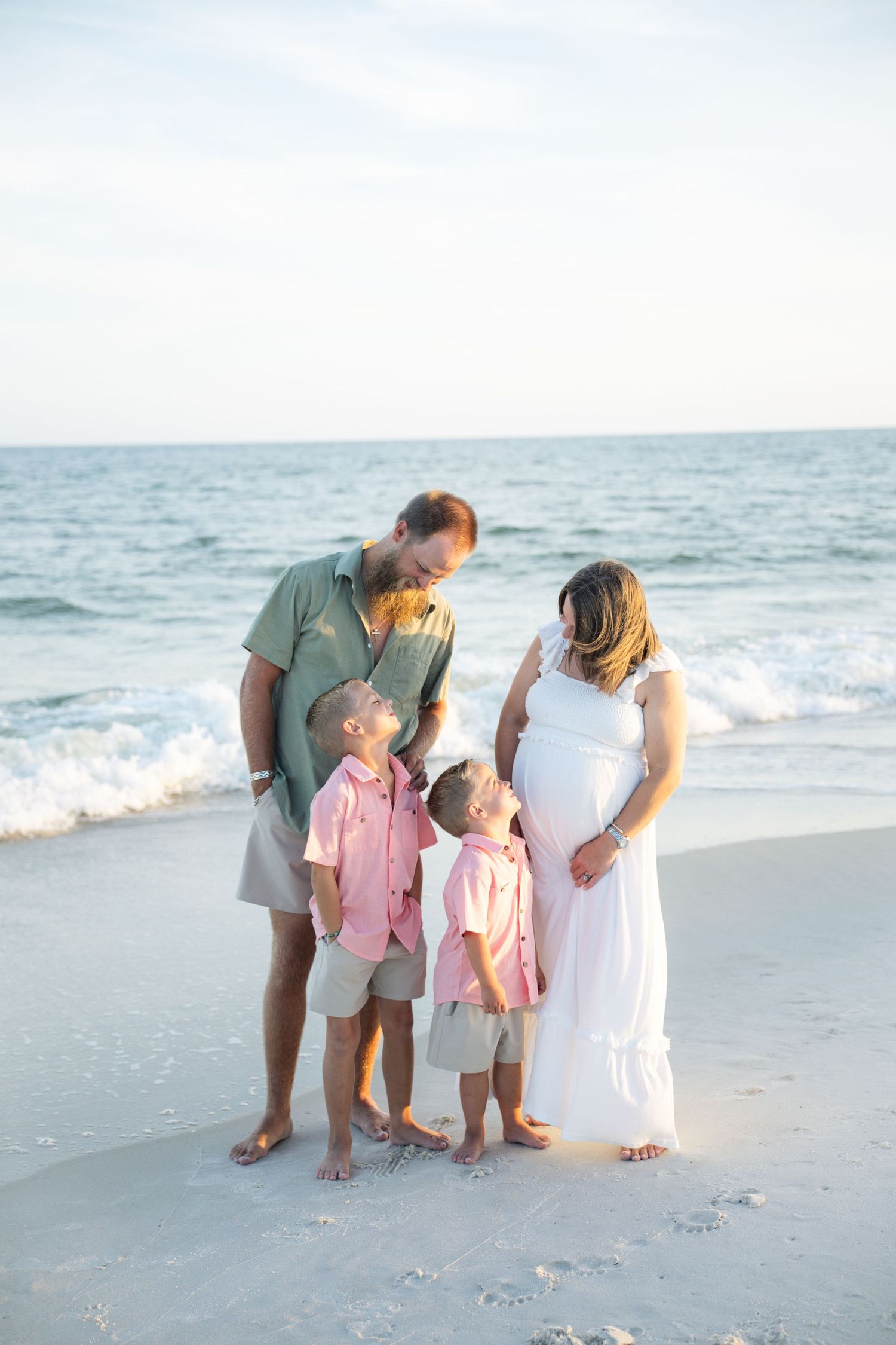 Pregnant woman in white dress with husband and two boys in pink shirts pose on sandy beach with ocean waves behind them.