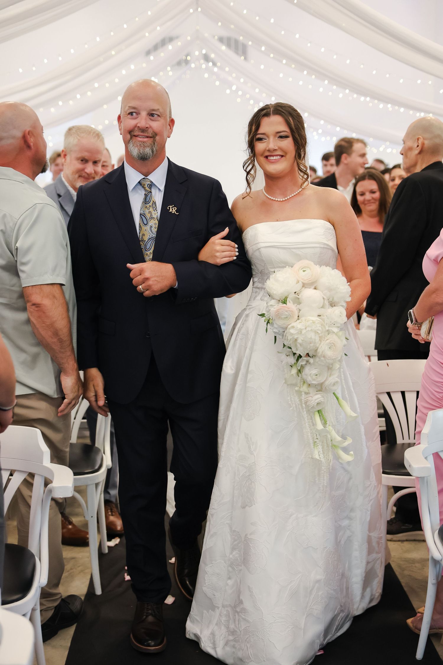 Bride in strapless white gown holding white bouquet walks down aisle with suited escort at elegant tent wedding.