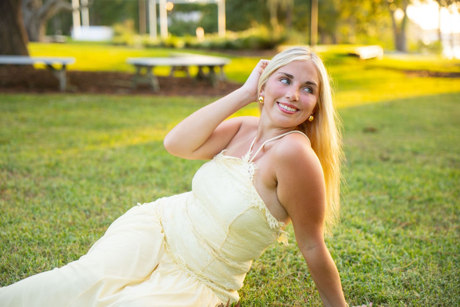Smiling blonde woman in elegant white dress sitting on green grass outdoors at golden hour.