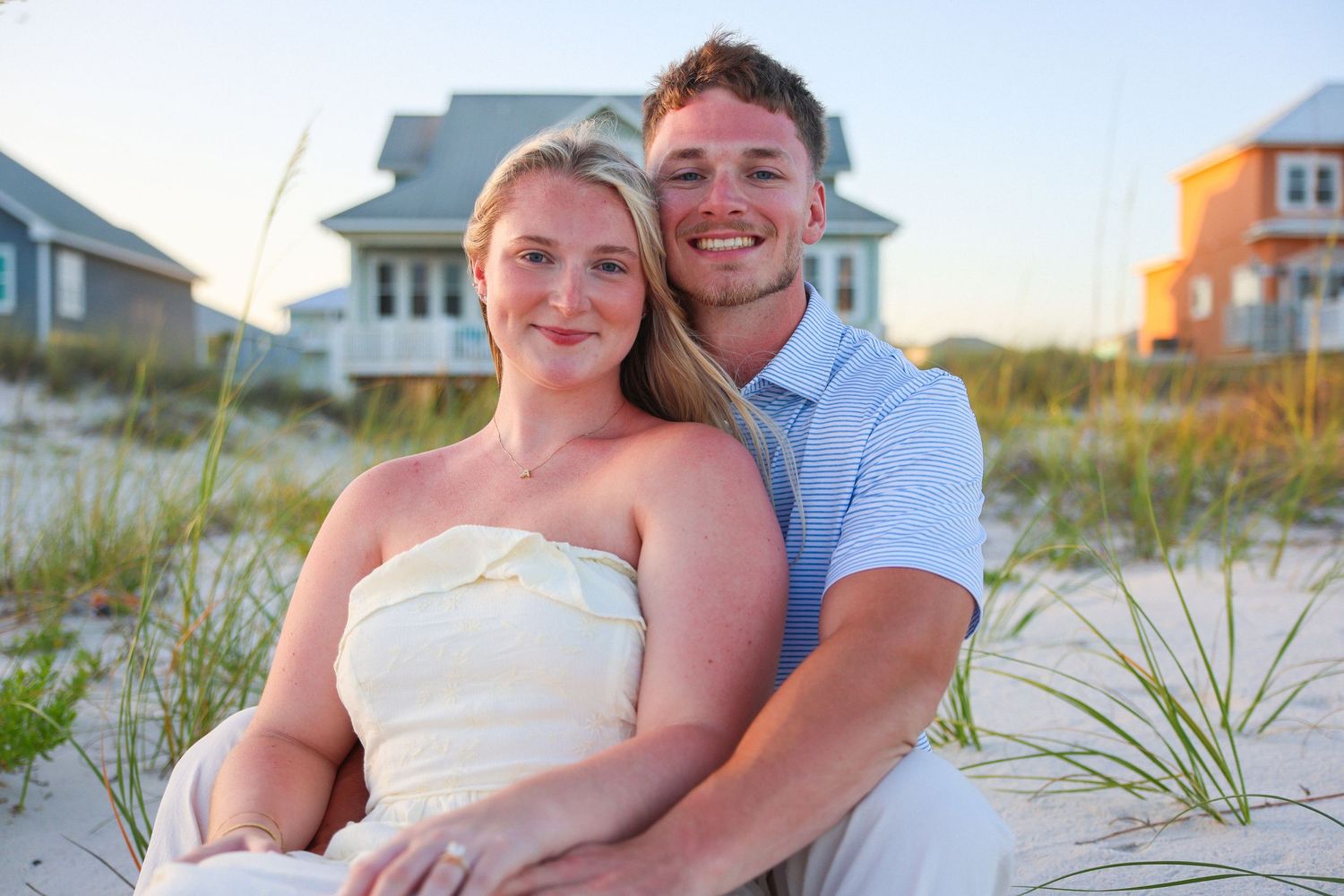 Happy couple smiling together on a sandy beach with coastal homes in the background on a sunny day.