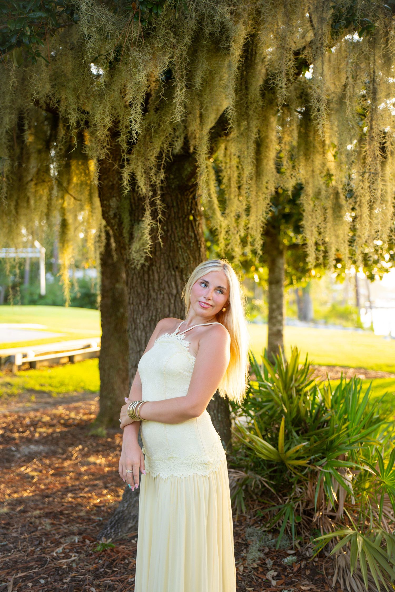 Blonde woman in yellow maxi dress standing under Spanish moss-draped tree in sunlit park setting.