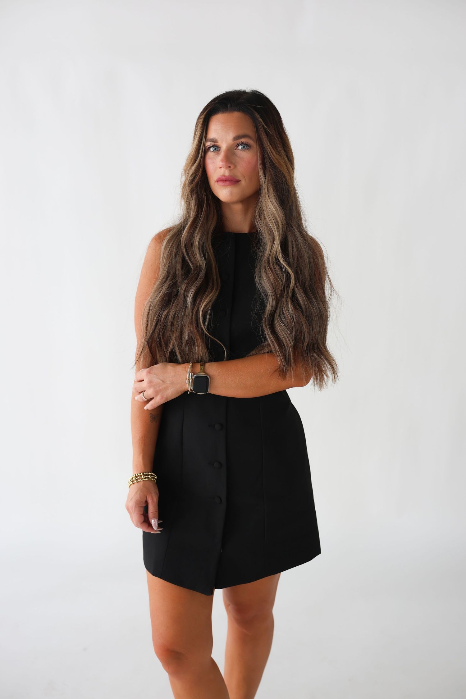 Woman in black sleeveless crop top and button-front mini skirt with long wavy ombre hair poses against white background.