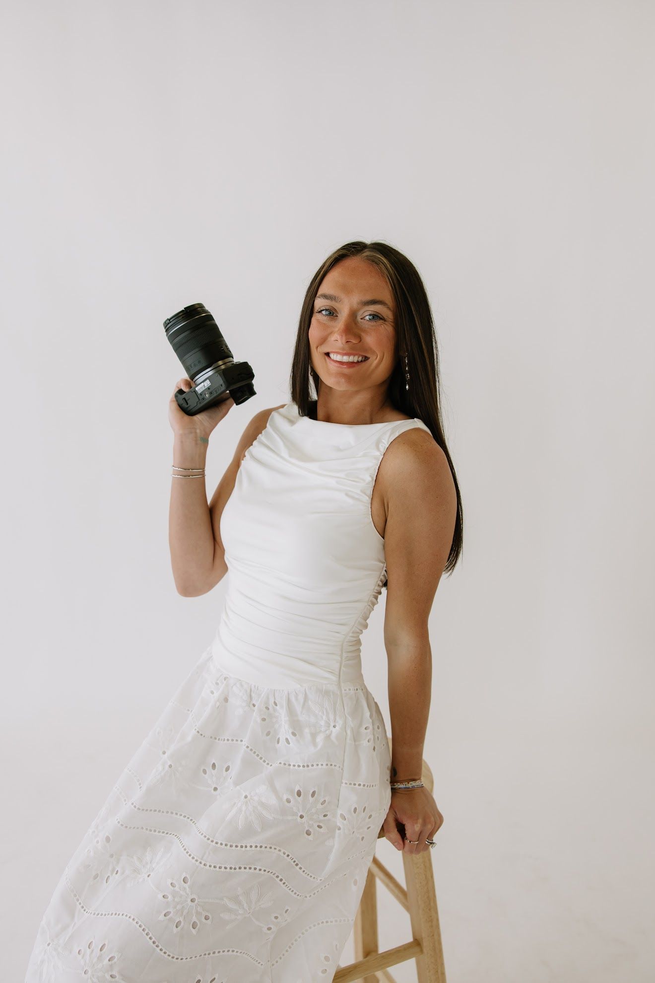 Smiling female photographer in white dress holding camera while leaning on stool.