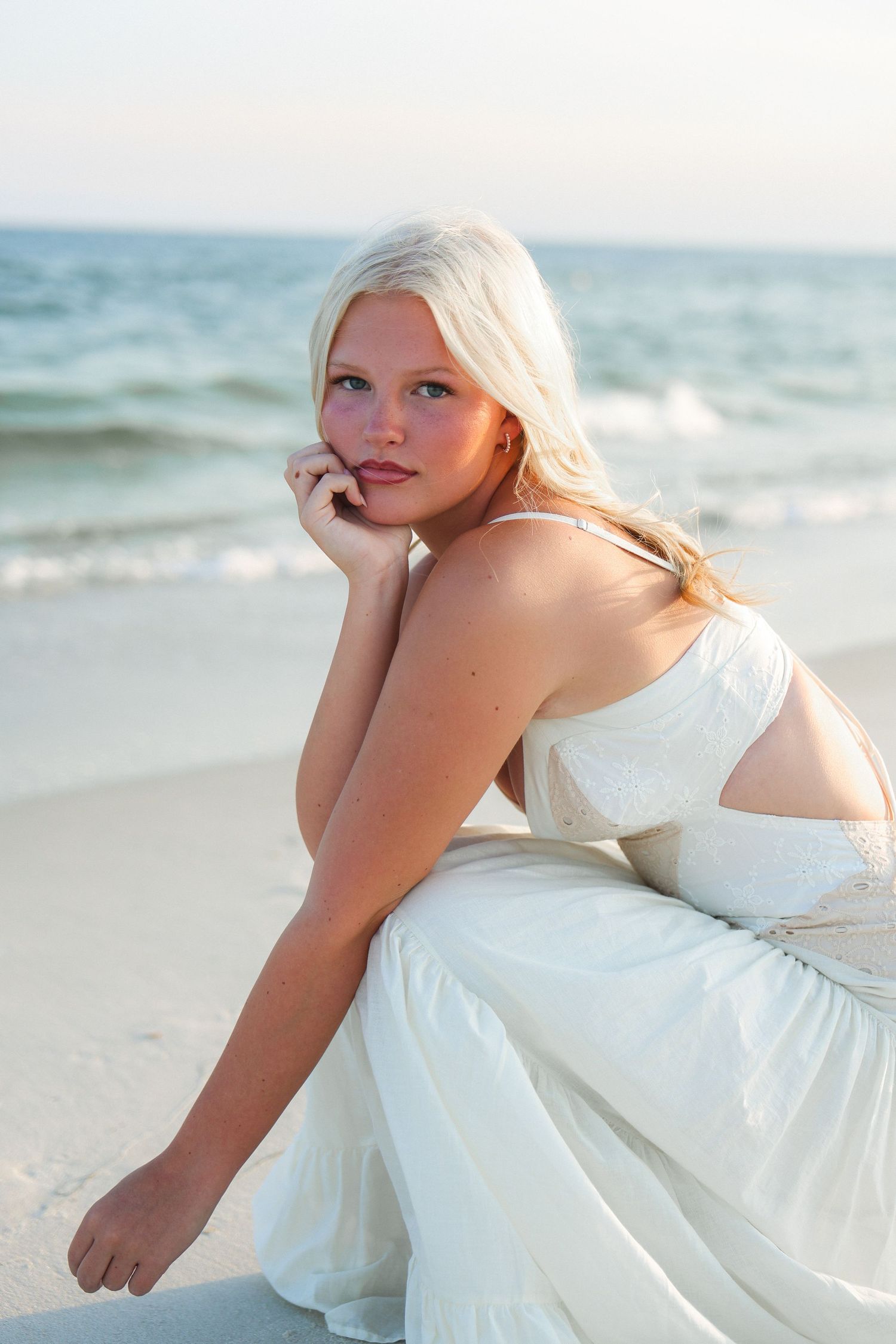 Young blonde woman in white dress sitting on sandy beach with ocean waves in background at sunset.