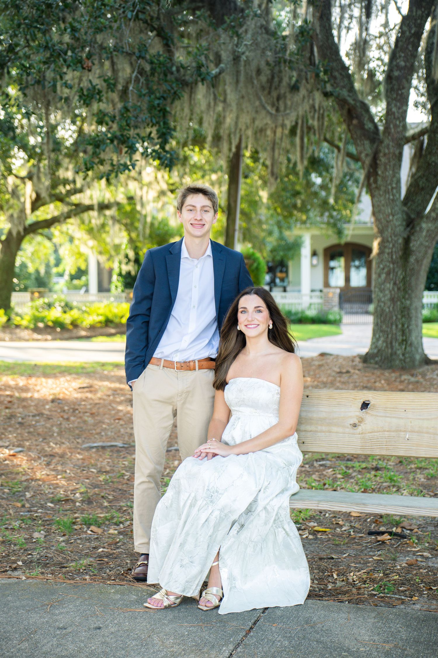 A young couple poses outdoors on a bench, woman in white dress, man in navy blazer, under moss-draped oak trees.