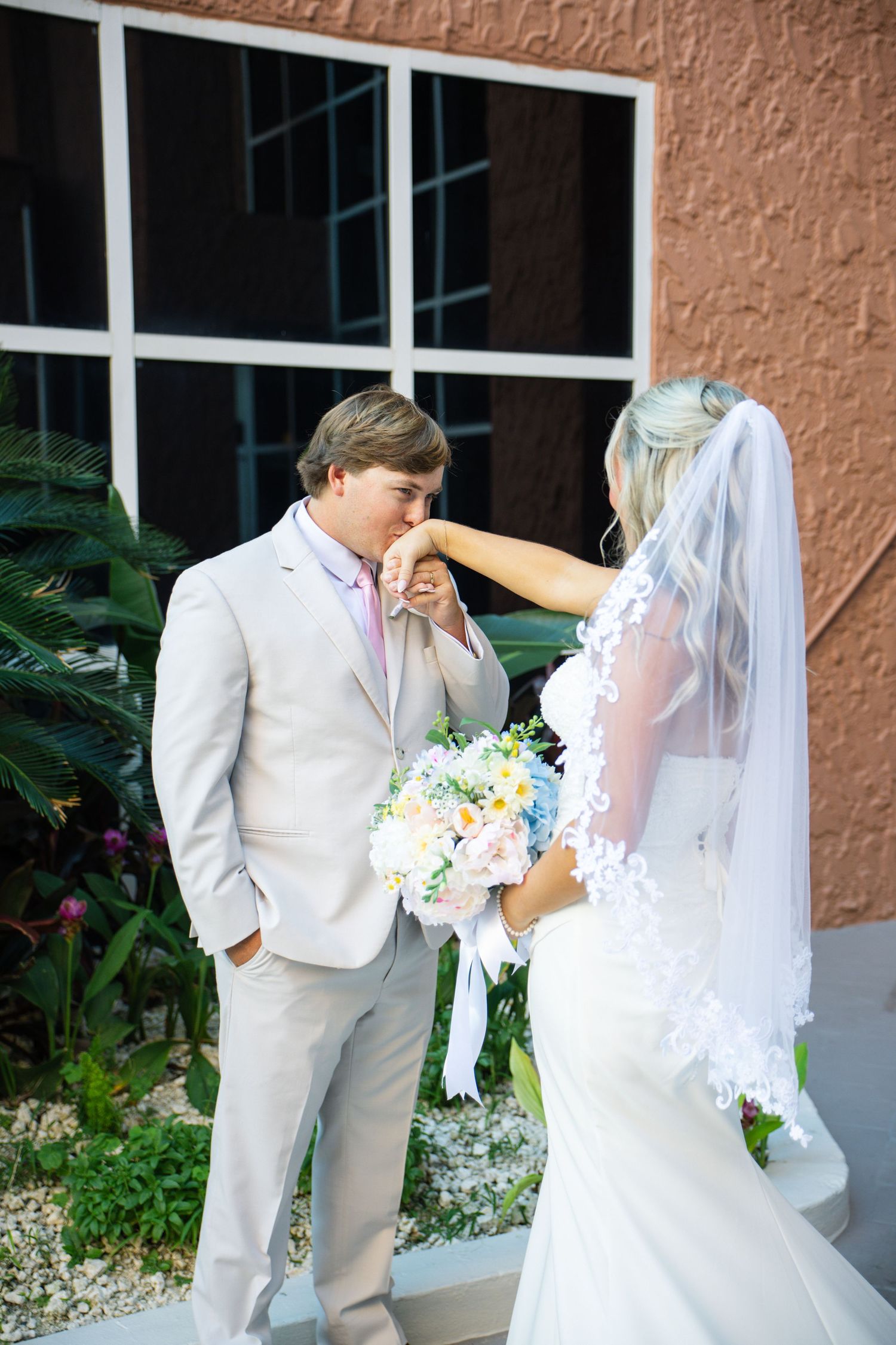 Groom in tan suit kisses bride's hand as she holds a pastel bouquet, wearing a white lace wedding dress and veil.