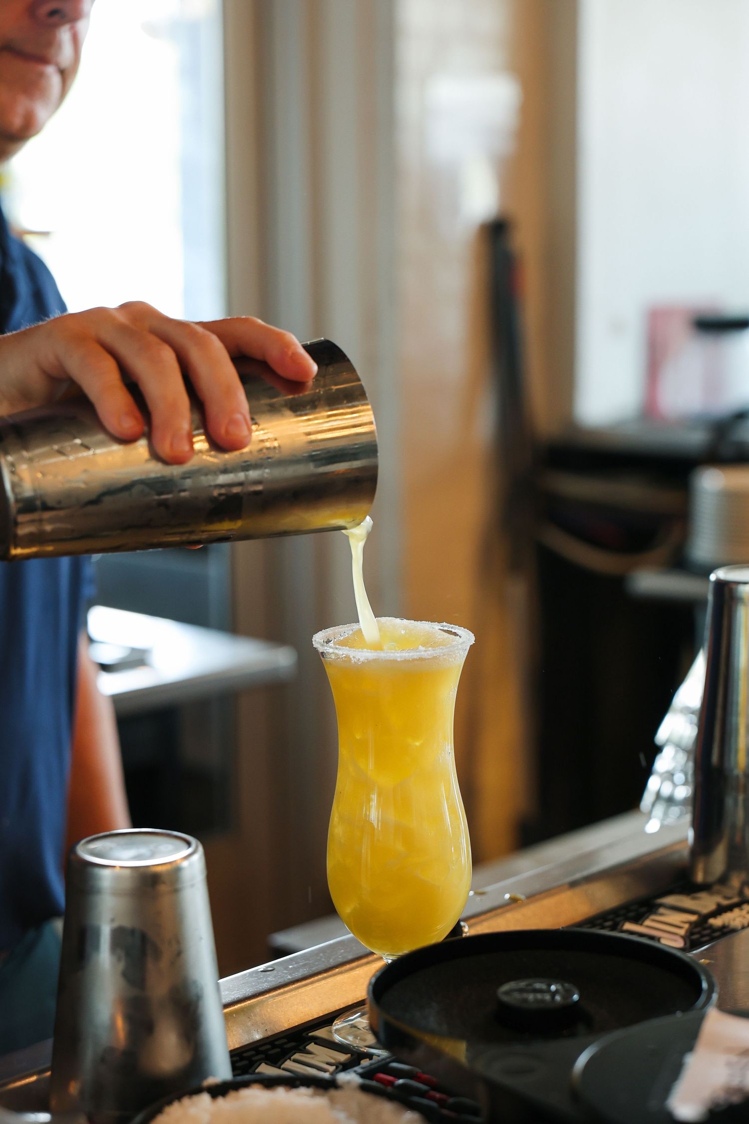 Bartender pouring a frothy yellow cocktail from a shaker into a hurricane glass at a bar.