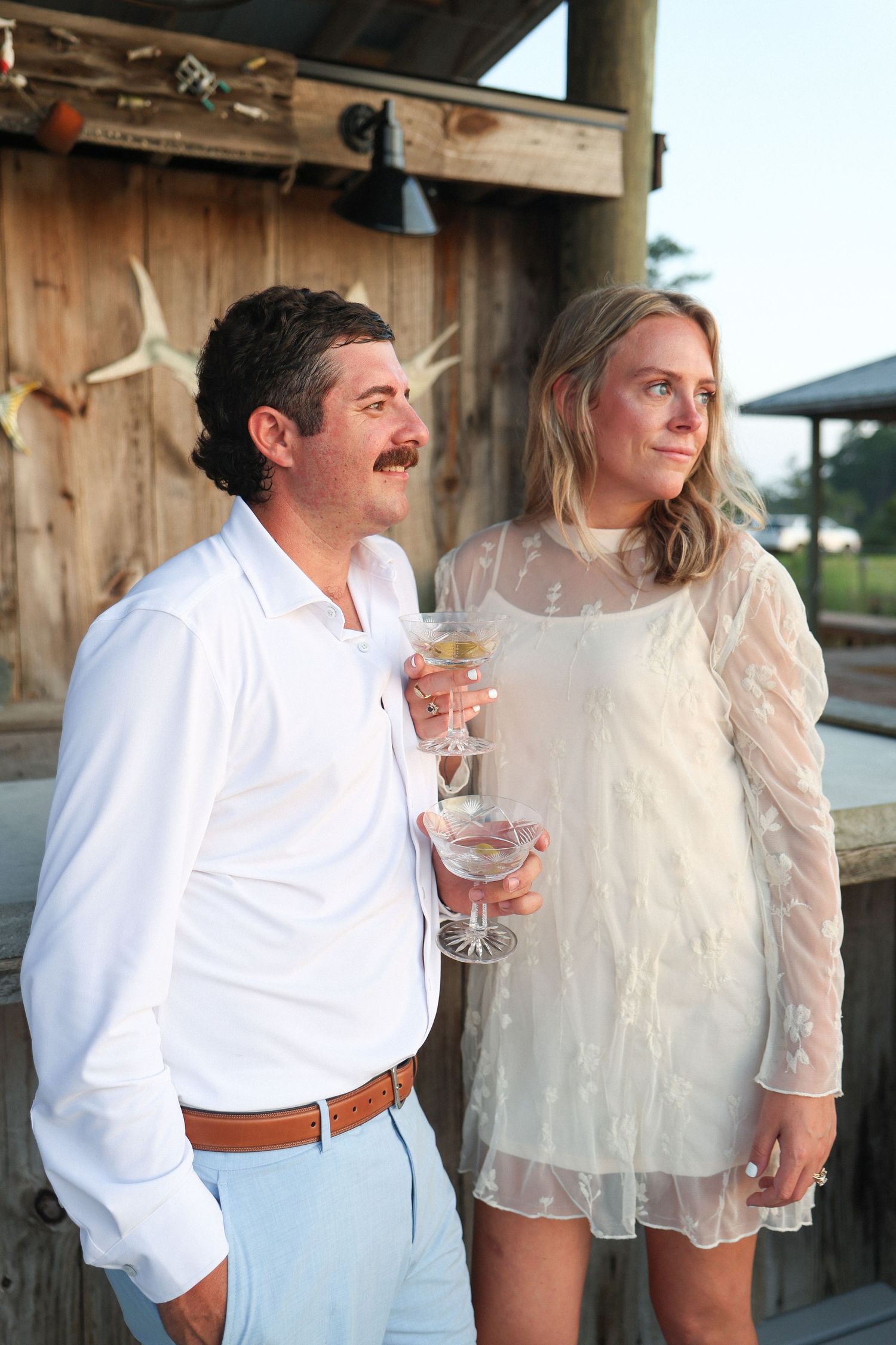 Couple in white attire holding wine glasses at an outdoor rustic venue during golden hour.