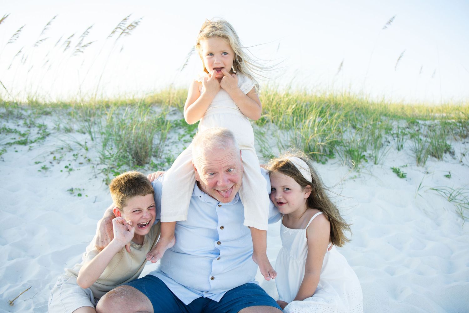 Grandfather sits on beach sand with three grandchildren, smiling and laughing together near sea oats on a sunny day.
