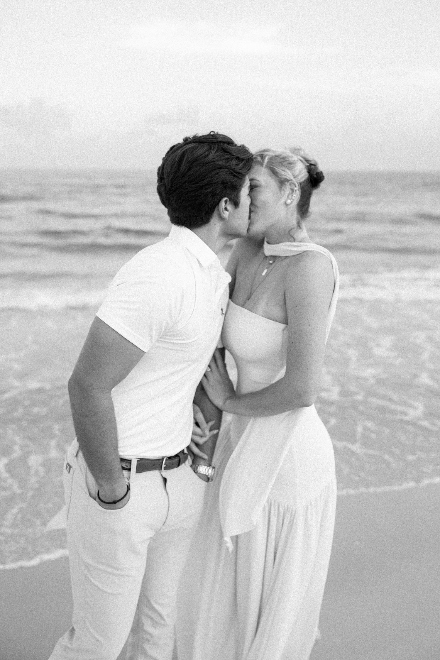 Black and white photo of a couple kissing on the beach, both dressed in white, with ocean waves in the background.