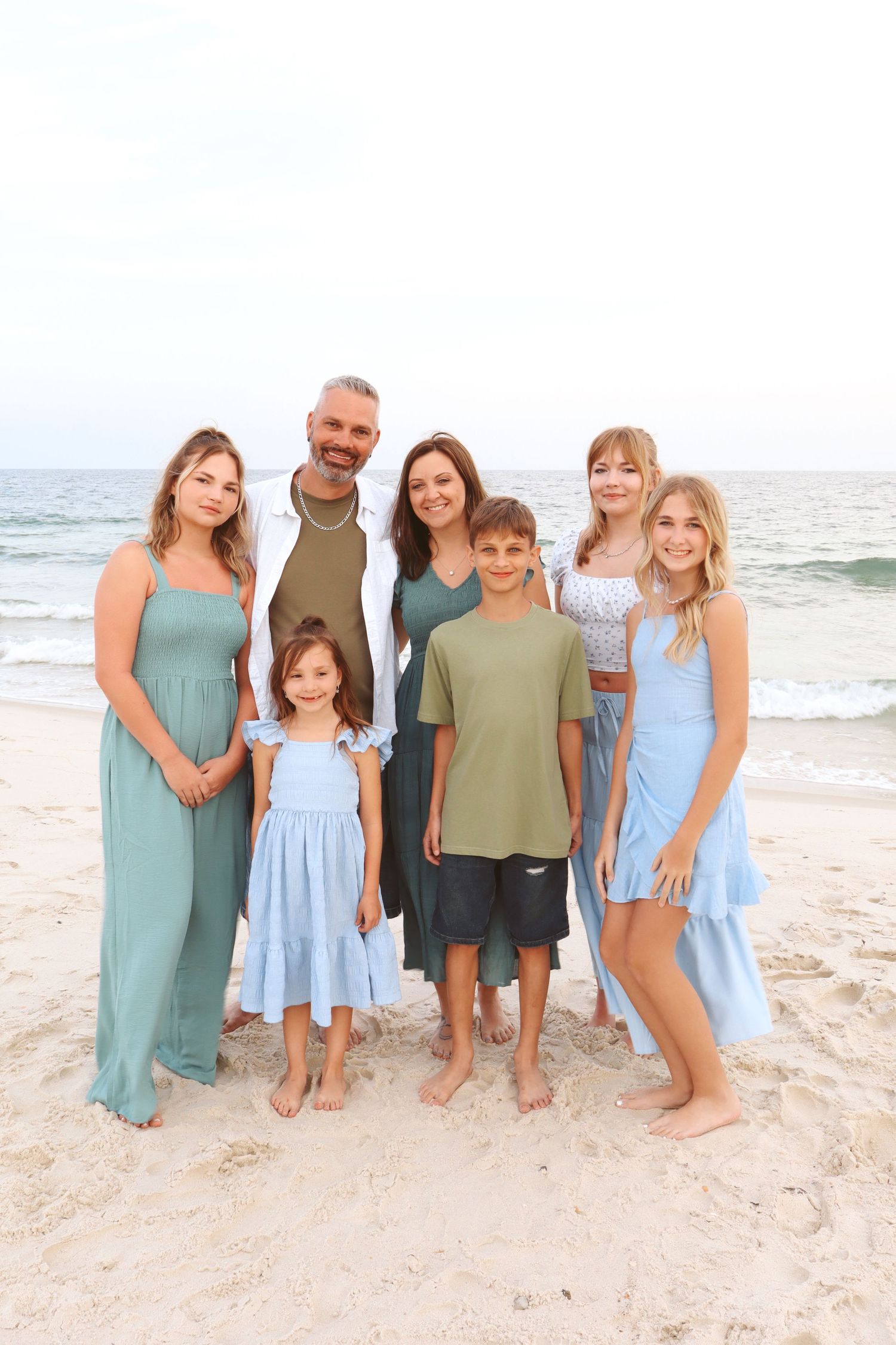 Family of seven posing on beach in coordinated green and blue outfits with ocean waves behind them.