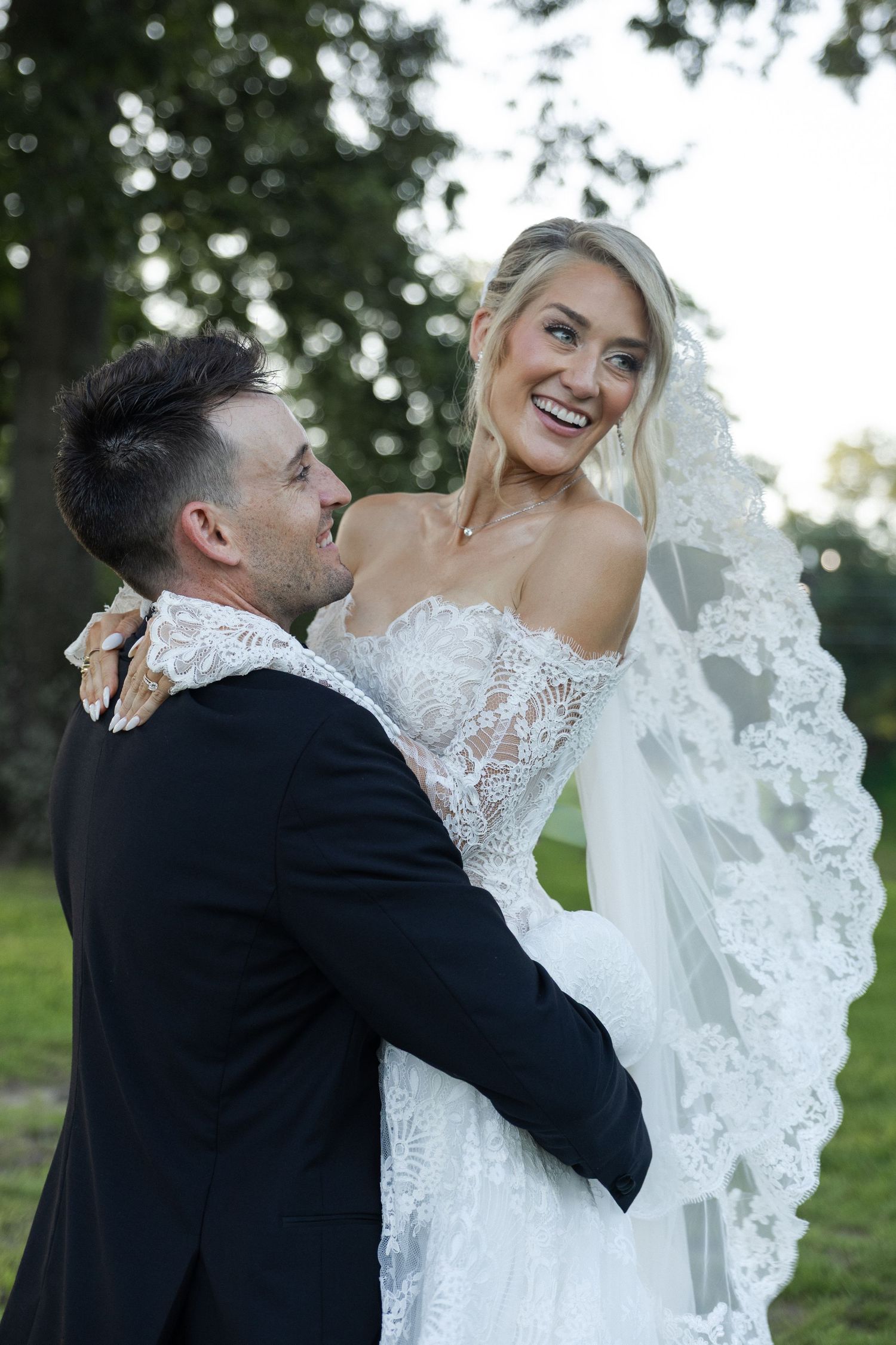 A joyful bride in a lace wedding gown and veil laughs as her groom in a black suit embraces her outdoors.