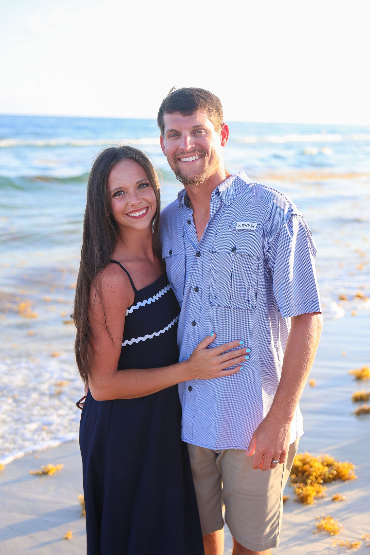 Smiling couple posing together on a sunny beach, woman in black dress and man in blue shirt, with ocean waves in background.