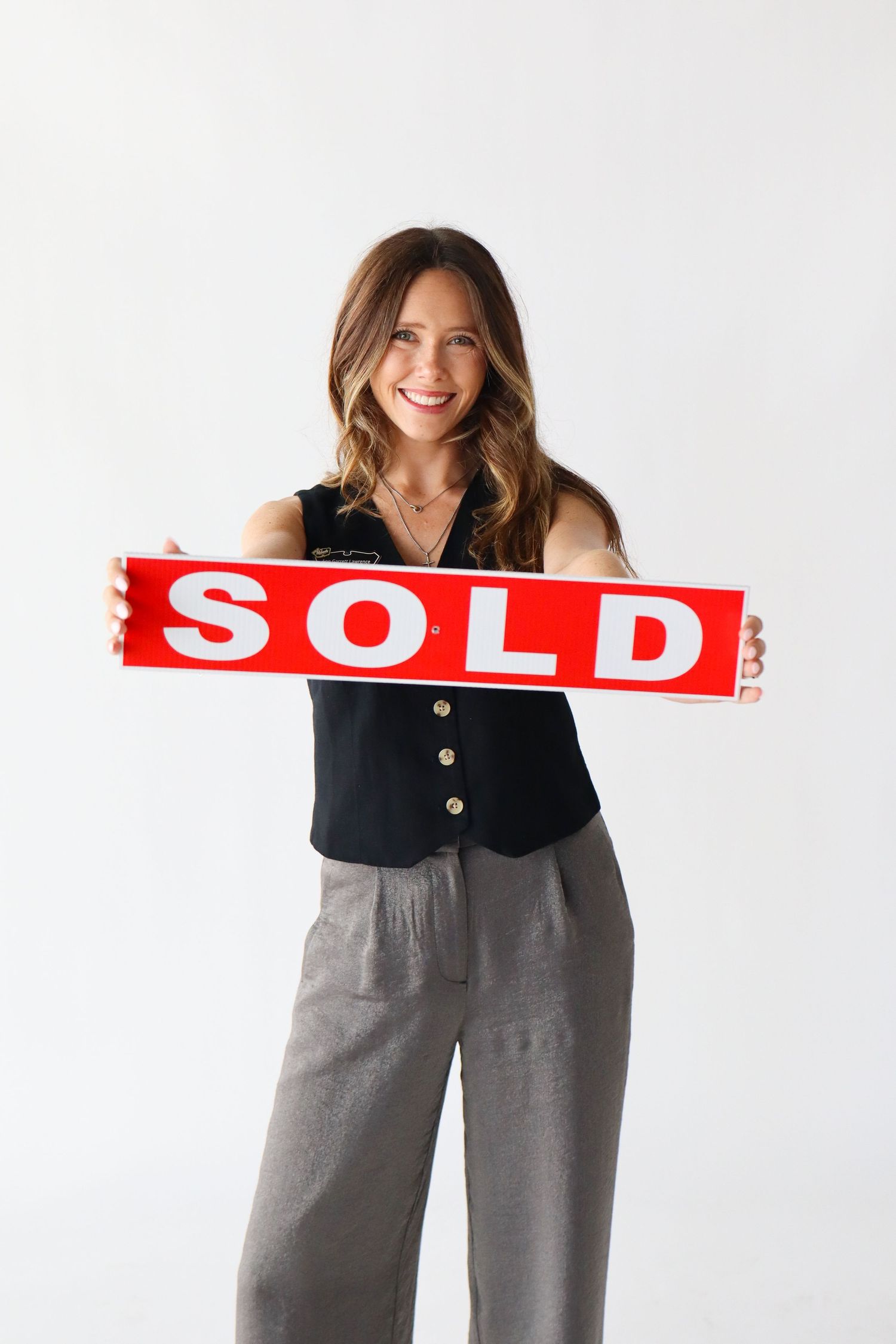 Smiling woman holding a red SOLD sign, wearing black vest and gray pants against white background.