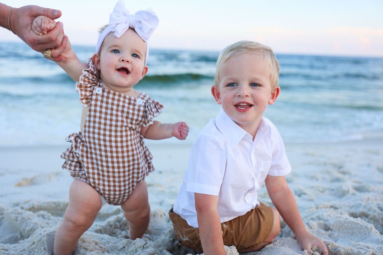 Two adorable toddlers smiling on a sandy beach, baby girl in gingham romper and boy in white shirt.
