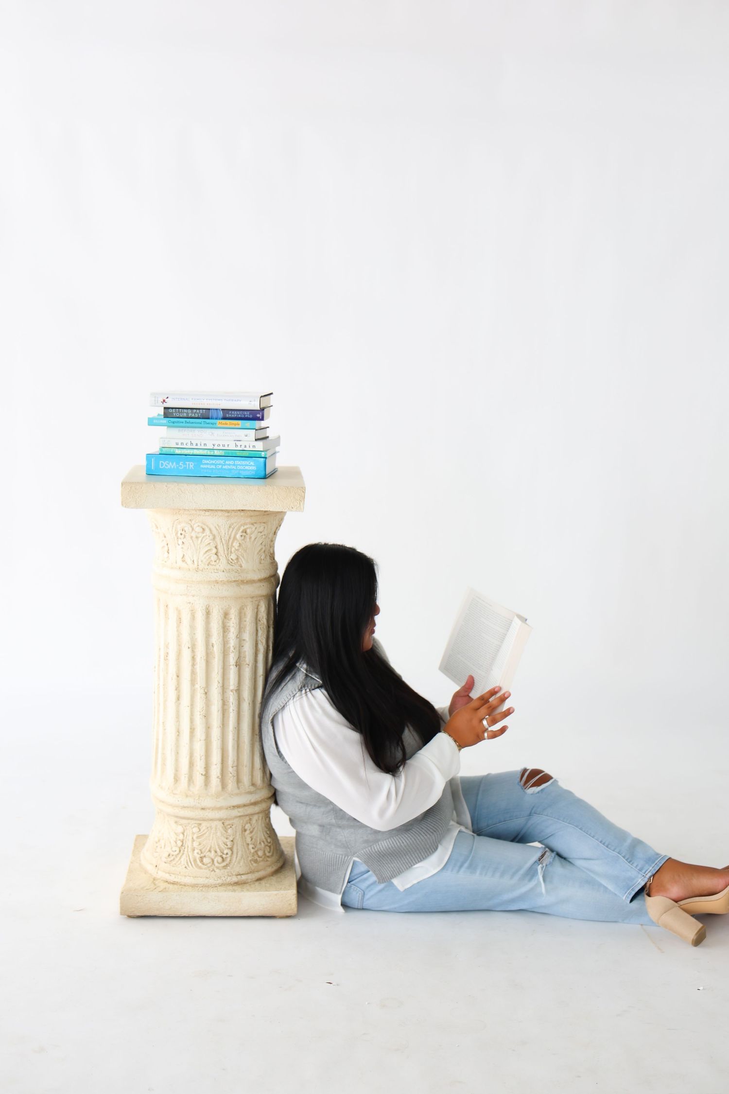 Young woman with long black hair sits on floor reading a book, leaning against a decorative column with stacked books.