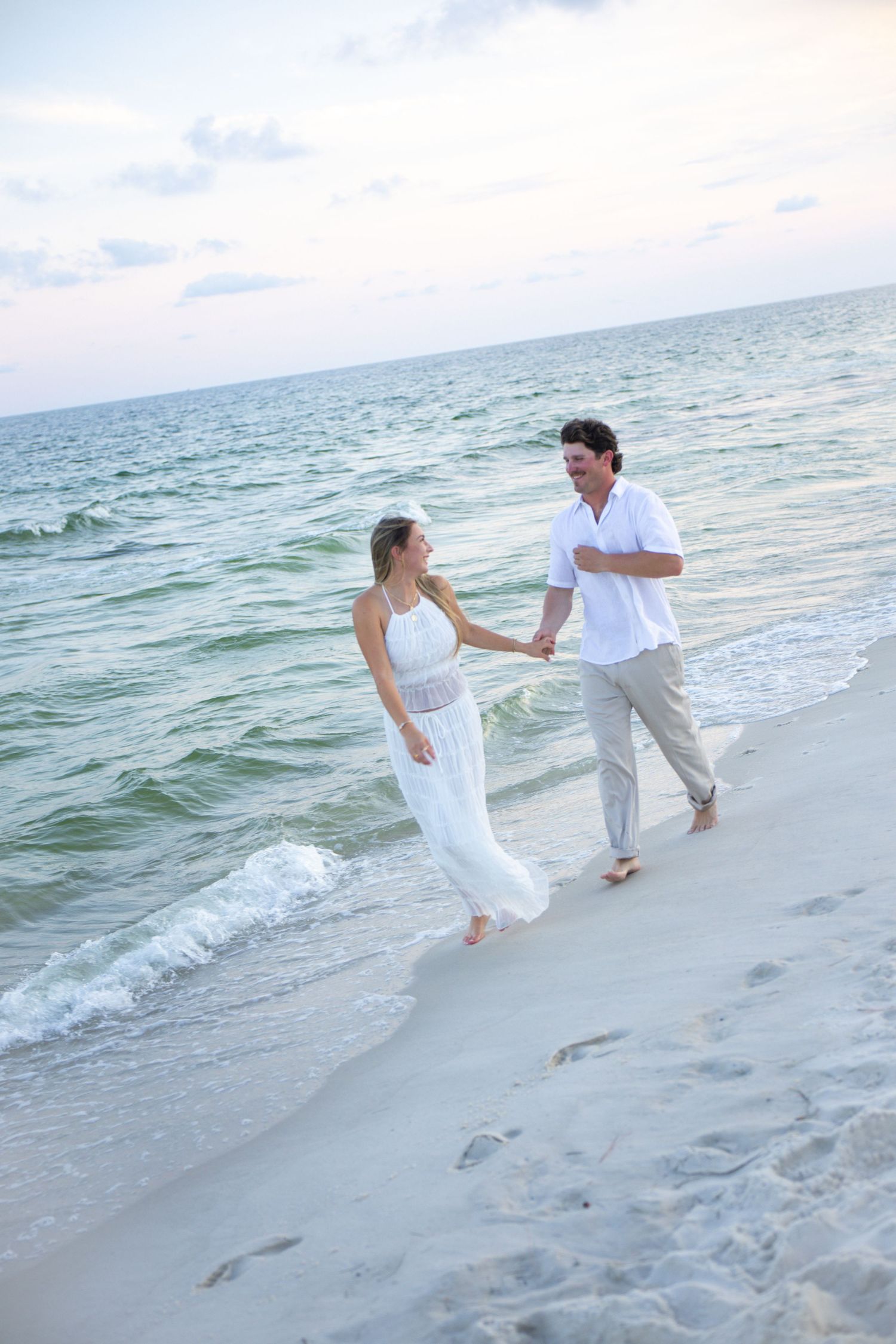 Couple in white outfits walking hand-in-hand along a serene beach at sunset, with gentle waves and footprints in the sand.