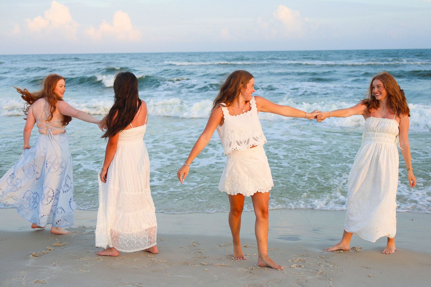Four girls in white dresses hold hands and dance together on a sandy beach with ocean waves in the background.