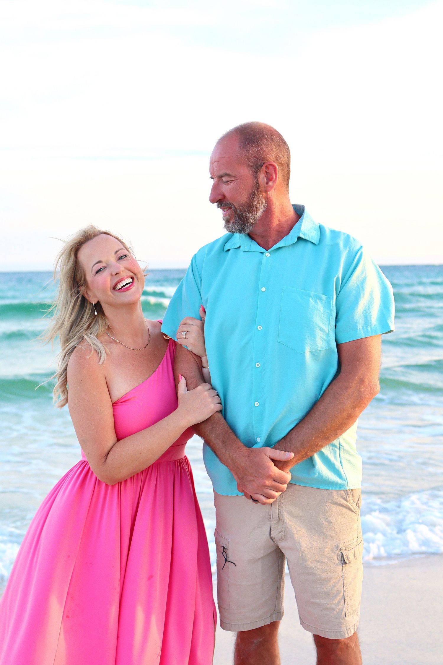 Laughing couple on beach, woman in pink dress holding mans arm in turquoise shirt near ocean waves.