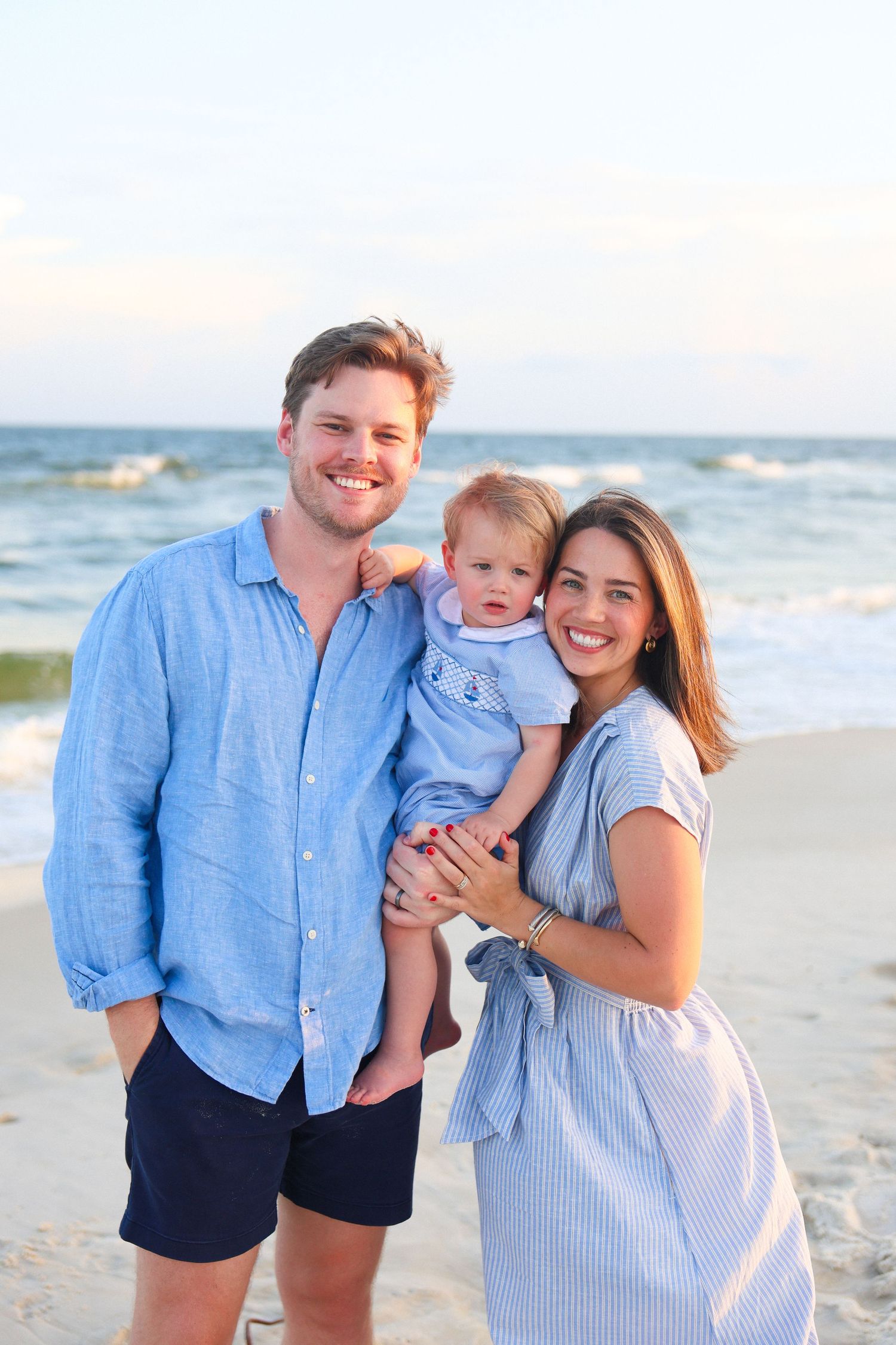 Happy young family with baby posing on sandy beach, wearing coordinating blue outfits with ocean waves behind them.