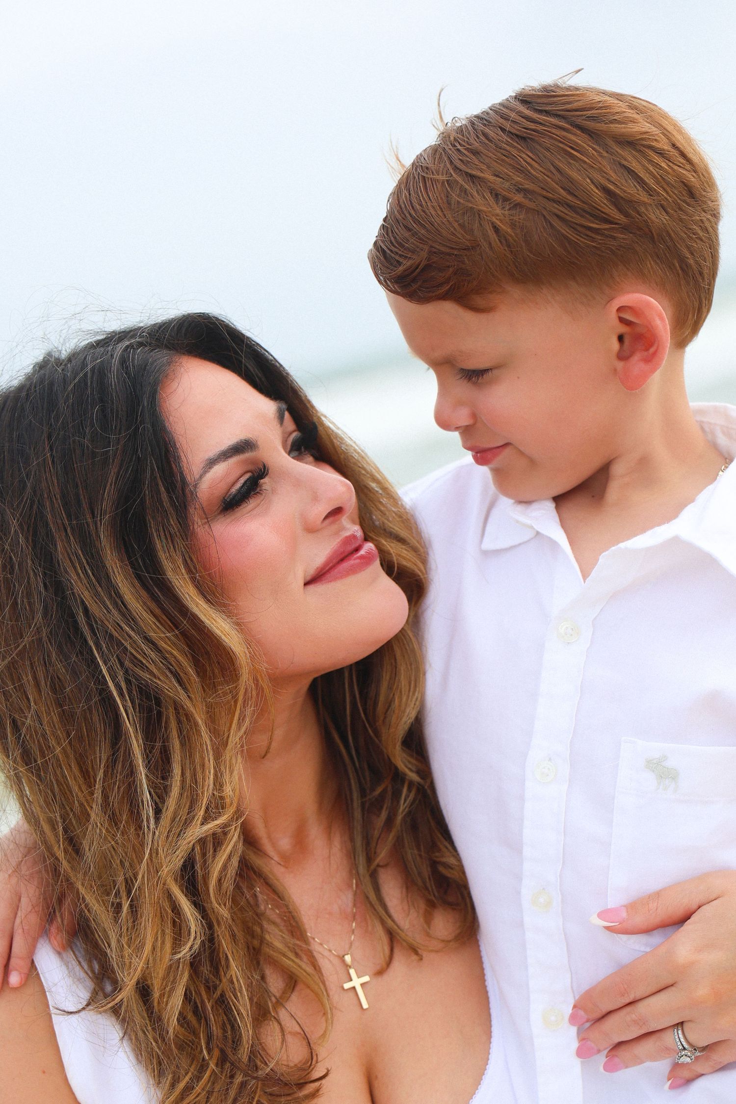 A woman with long brunette hair wearing a cross necklace smiles at a young boy in a white shirt outdoors.