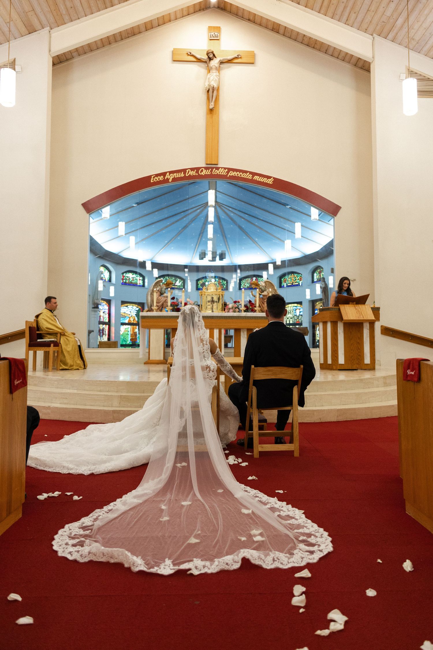 Bride and groom seated at Catholic church altar during wedding ceremony with long lace veil trailing on red carpet.