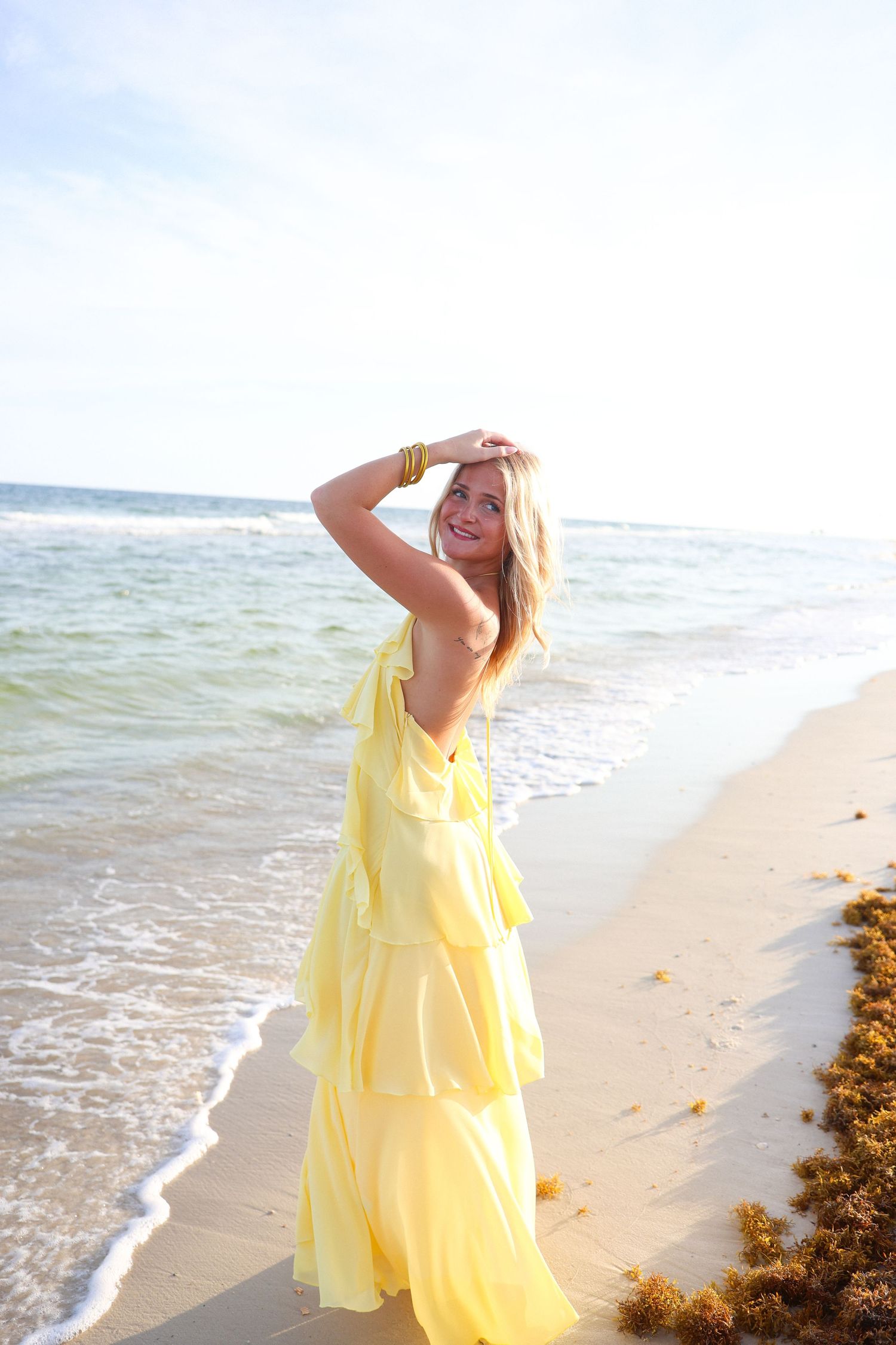 Smiling woman in yellow ruffled dress posing on sandy beach with ocean waves in background.