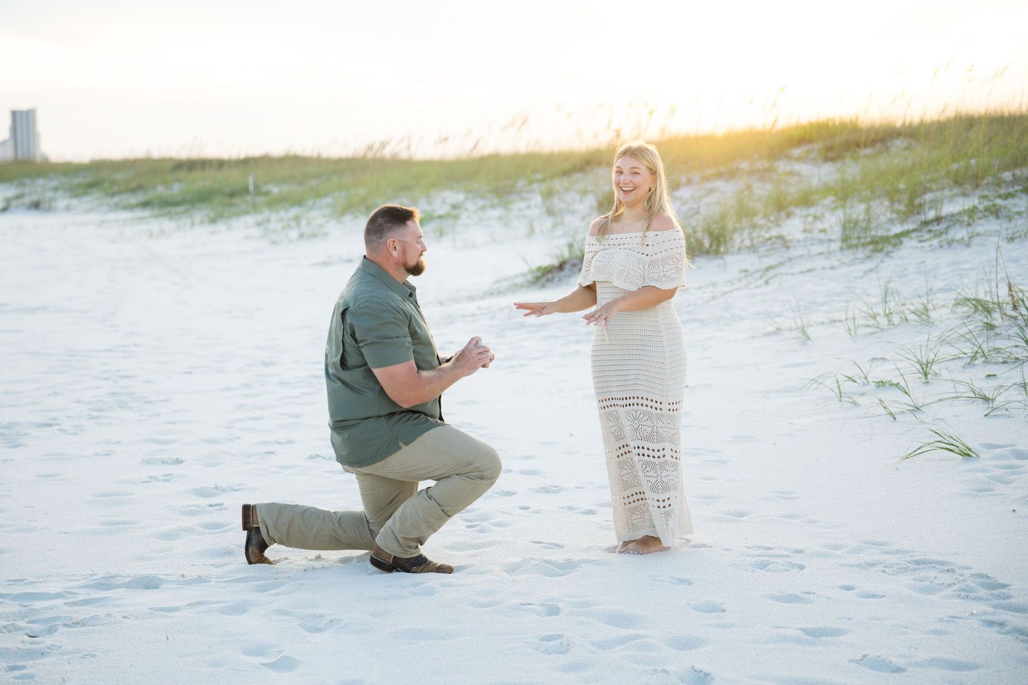Man proposing on one knee to woman in white dress on sandy beach at sunset.