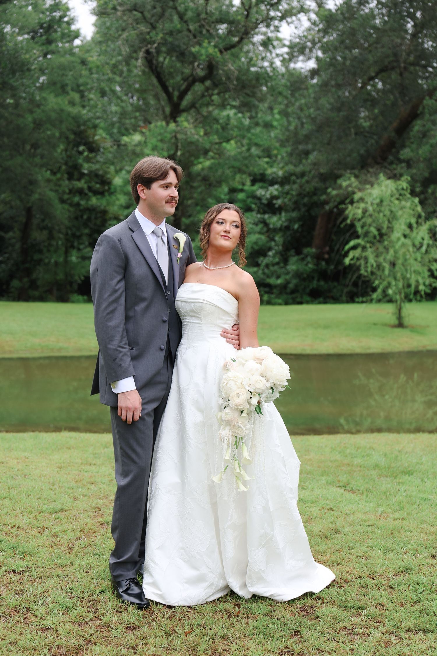 Bride in white strapless gown holding bouquet stands with groom in gray suit outdoors near pond.