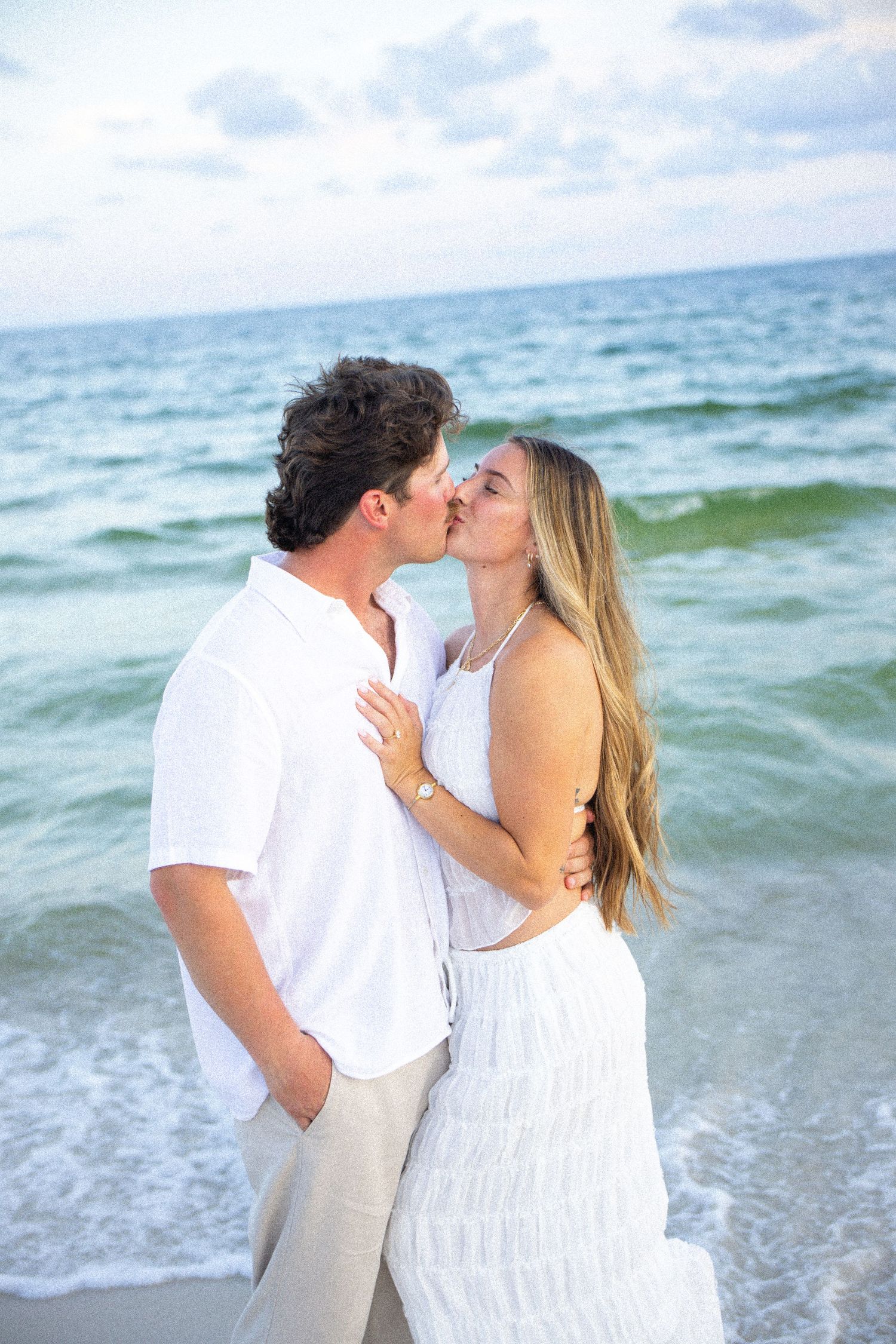 Couple sharing a romantic kiss on the beach, dressed in white outfits with ocean waves in the background.