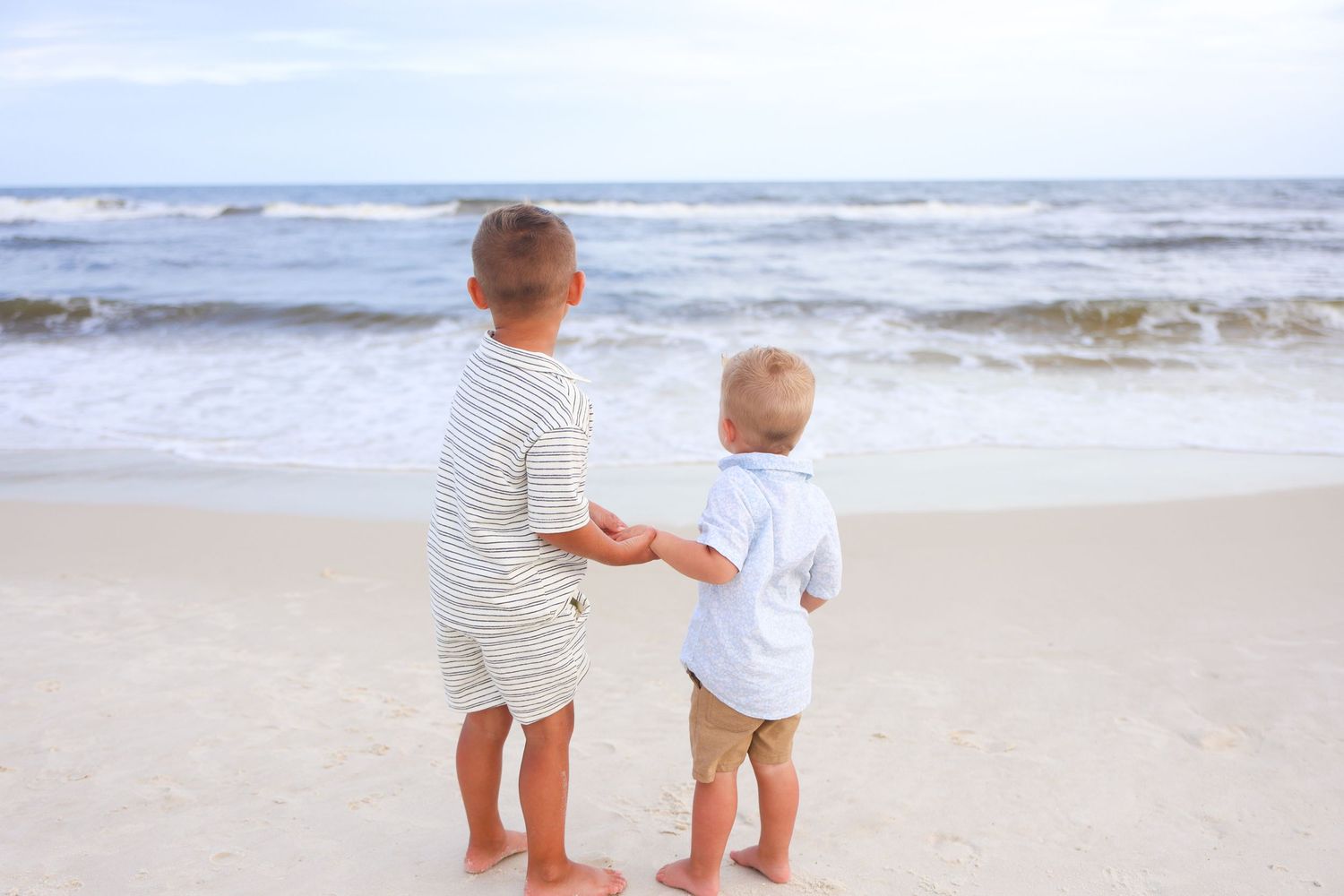 Two young boys standing barefoot on a sandy beach, holding hands while looking out at the ocean waves.