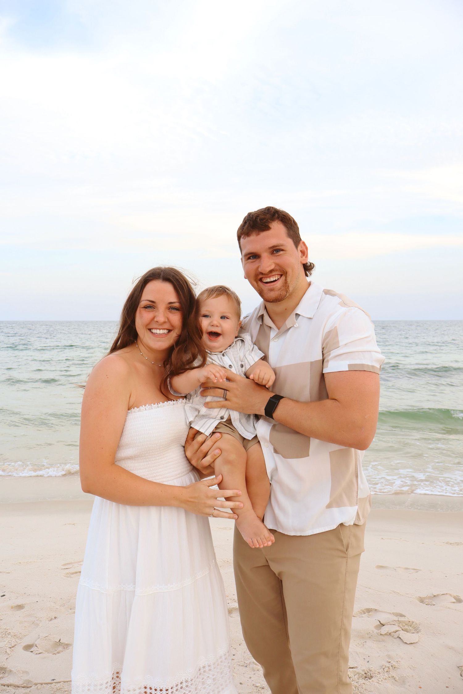 Smiling young family with laughing baby posing on a sunny beach in white and neutral outfits.