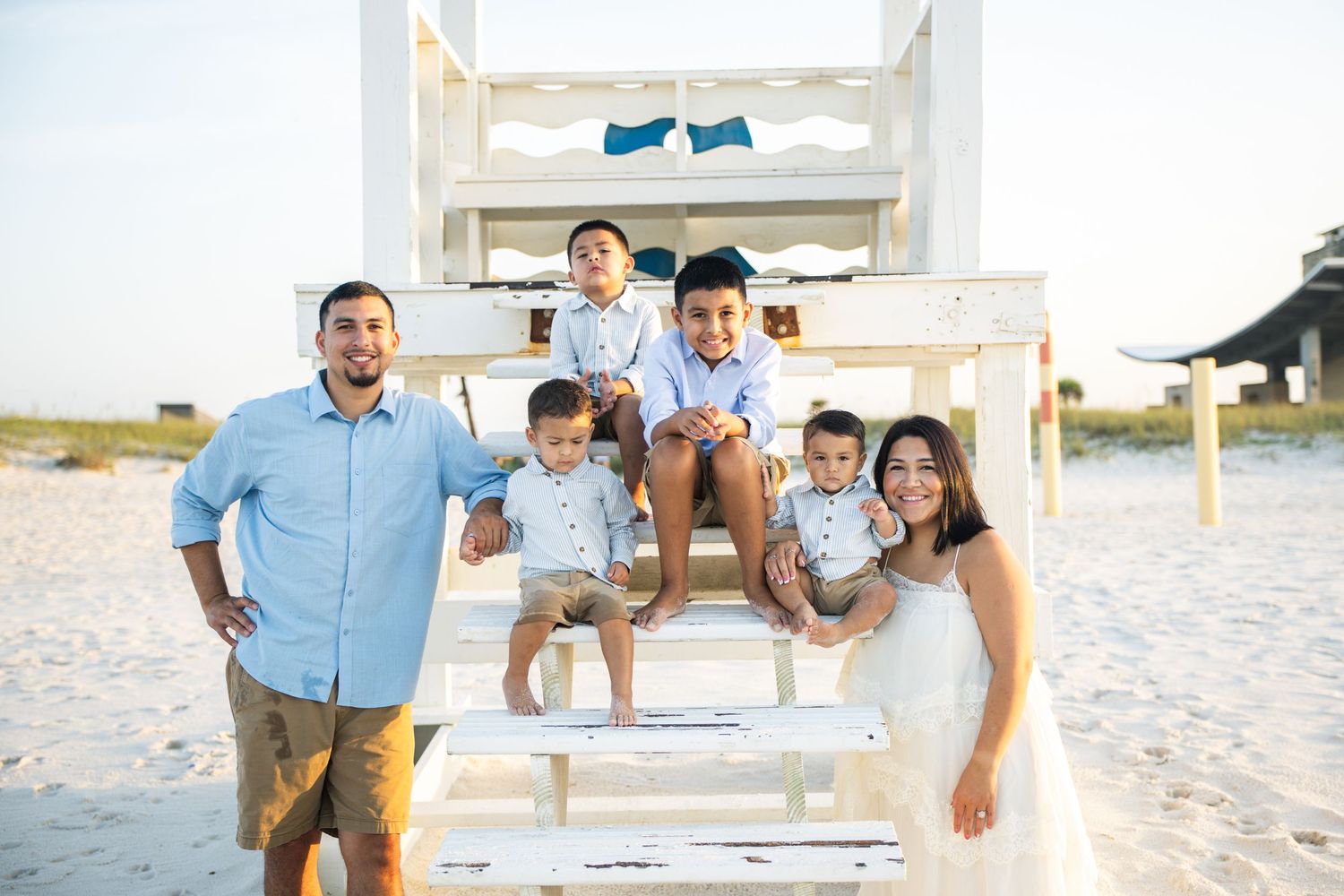 Family of six posing by a white lifeguard stand on a sunny beach, parents and four young boys in coordinating outfits.