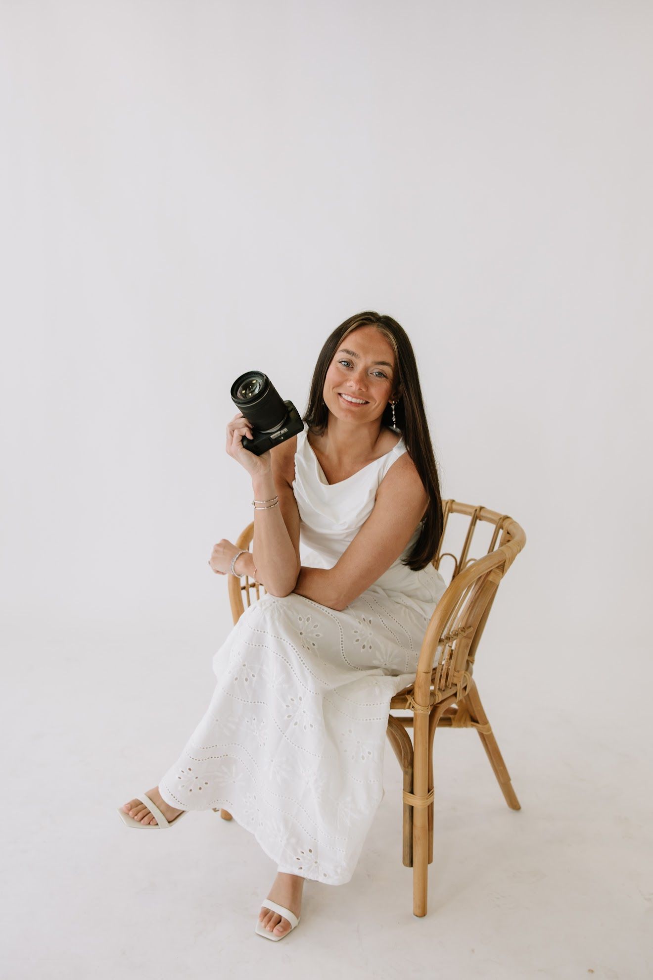 Woman in white dress sitting on wicker chair holding a professional camera, smiling in a bright studio.