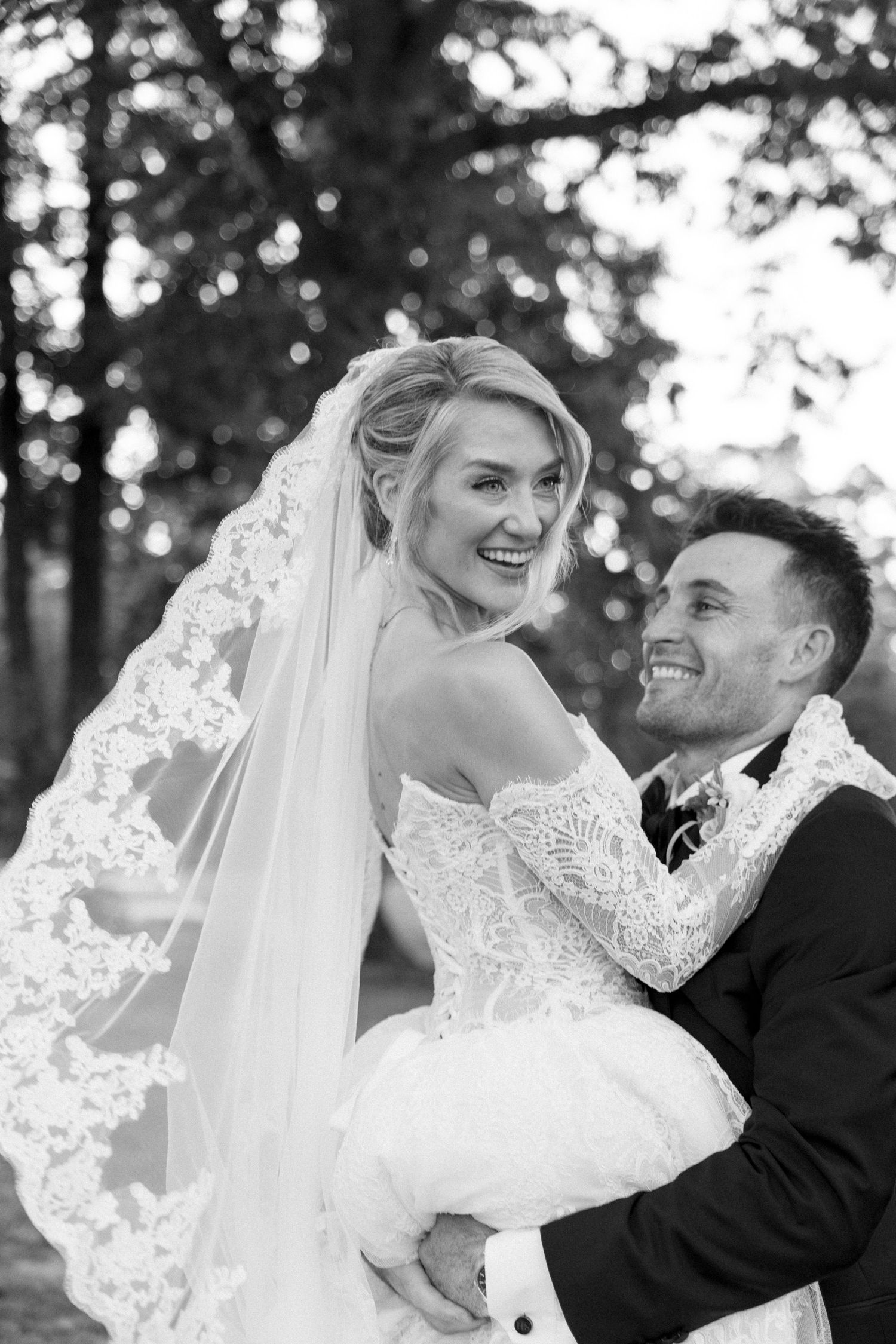 Black and white photo of a smiling bride in lace gown and veil being lifted by a groom in a suit outdoors.
