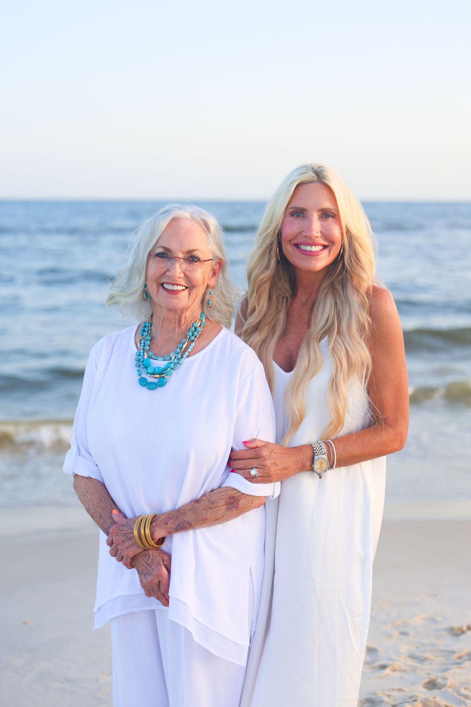 Two women in white outfits smiling on a sunny beach, one older with turquoise jewelry, one younger with long blonde hair.