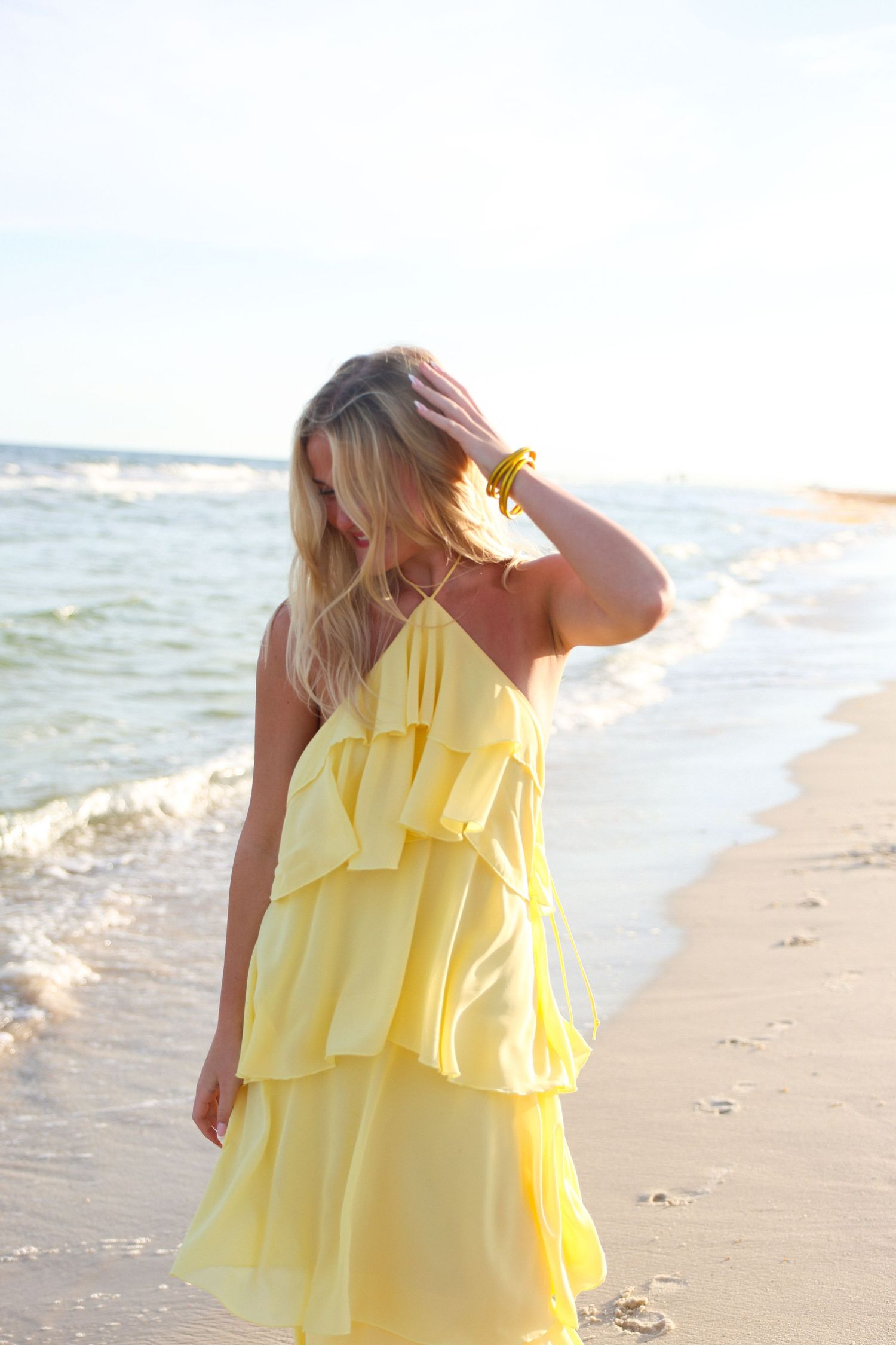 Woman in yellow ruffled sundress standing on sandy beach with ocean waves, hand in blonde hair.