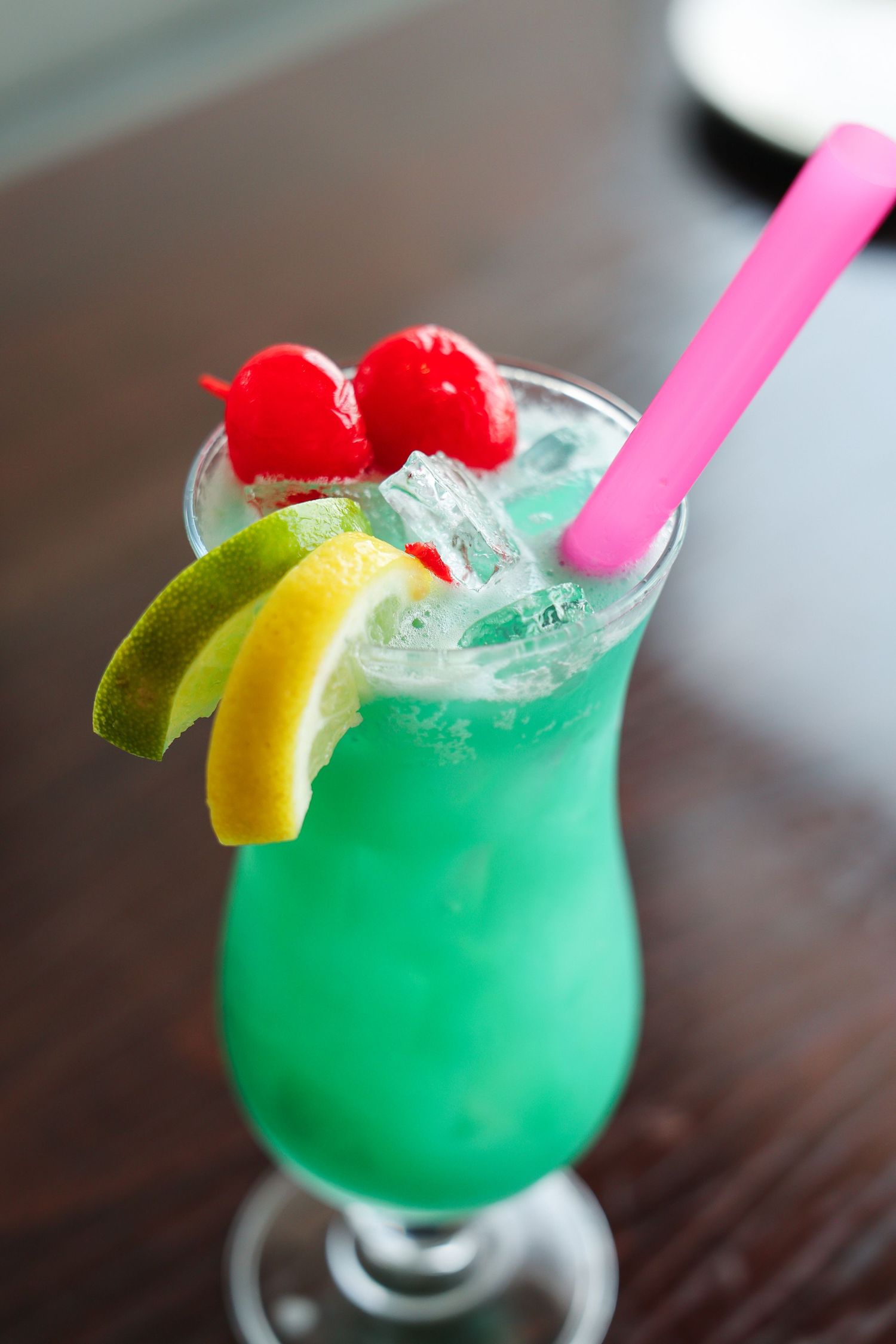 Vibrant green tropical cocktail with cherries, lime, lemon slices, ice, and a pink straw in a hurricane glass.