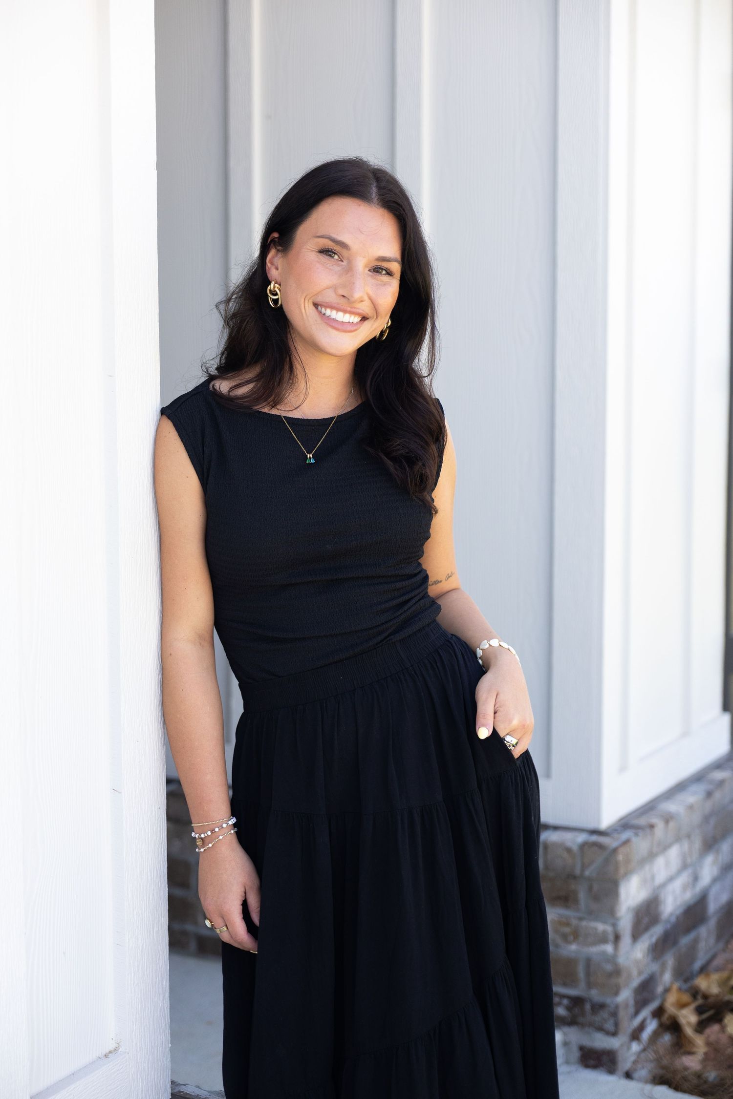 Smiling woman in black sleeveless dress leaning against white column outdoors.