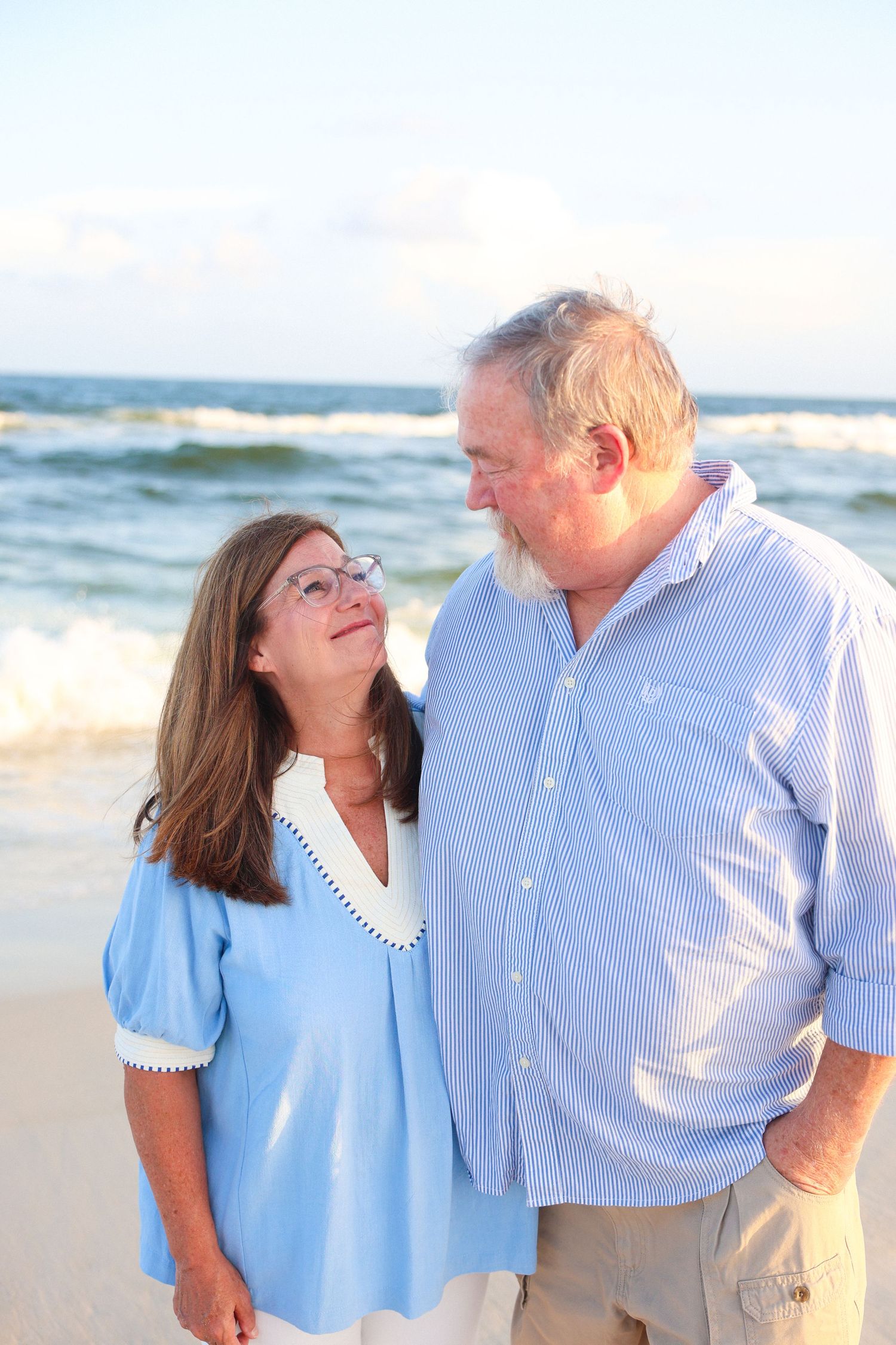 An elderly couple shares a tender moment on a sunny beach, the woman in blue gazing up at the man affectionately.