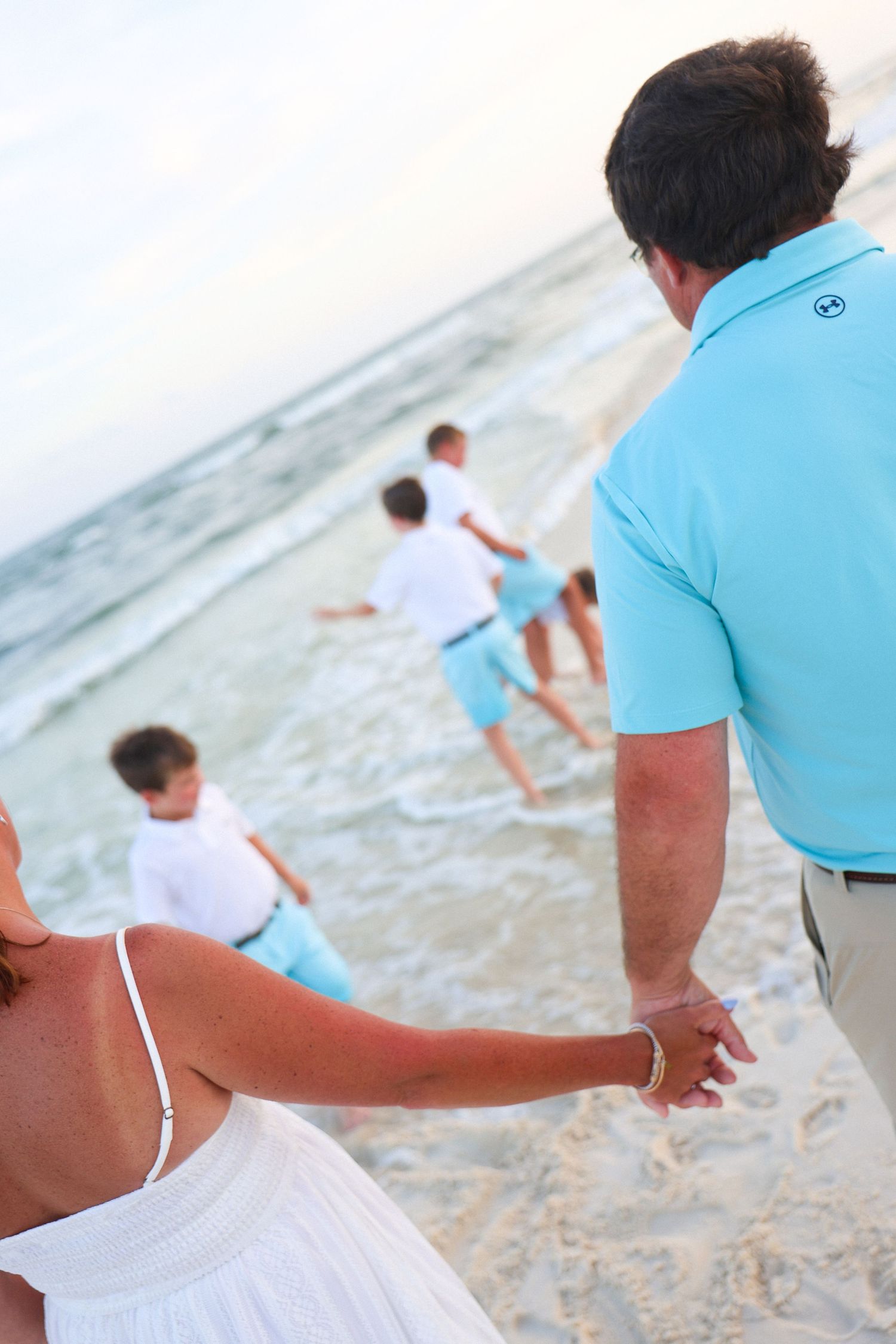 Family in matching white and turquoise outfits holding hands on a sandy beach at sunset.