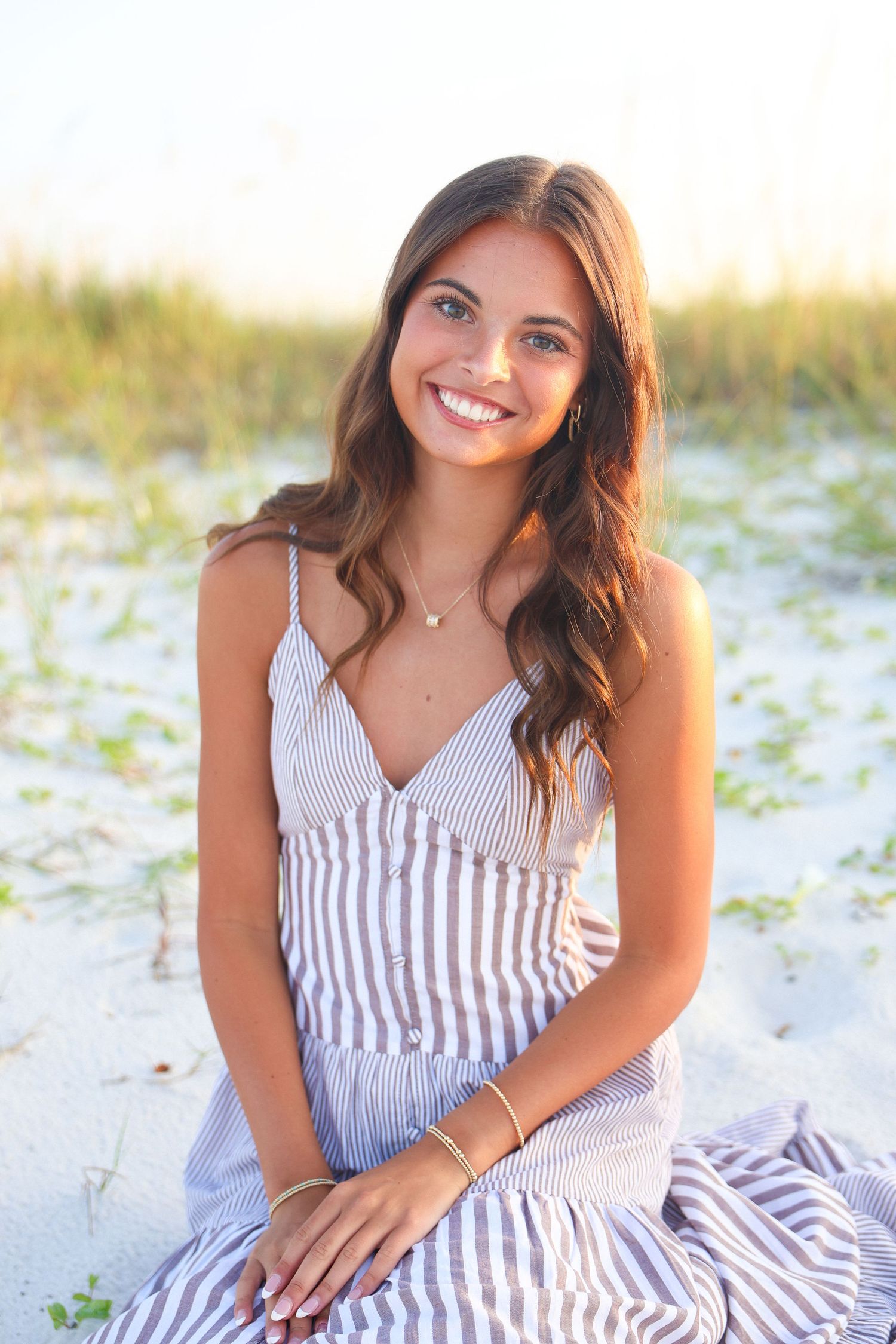 Young woman smiling on sandy beach, wearing striped sundress with gold jewelry, long brown hair, sunny day.