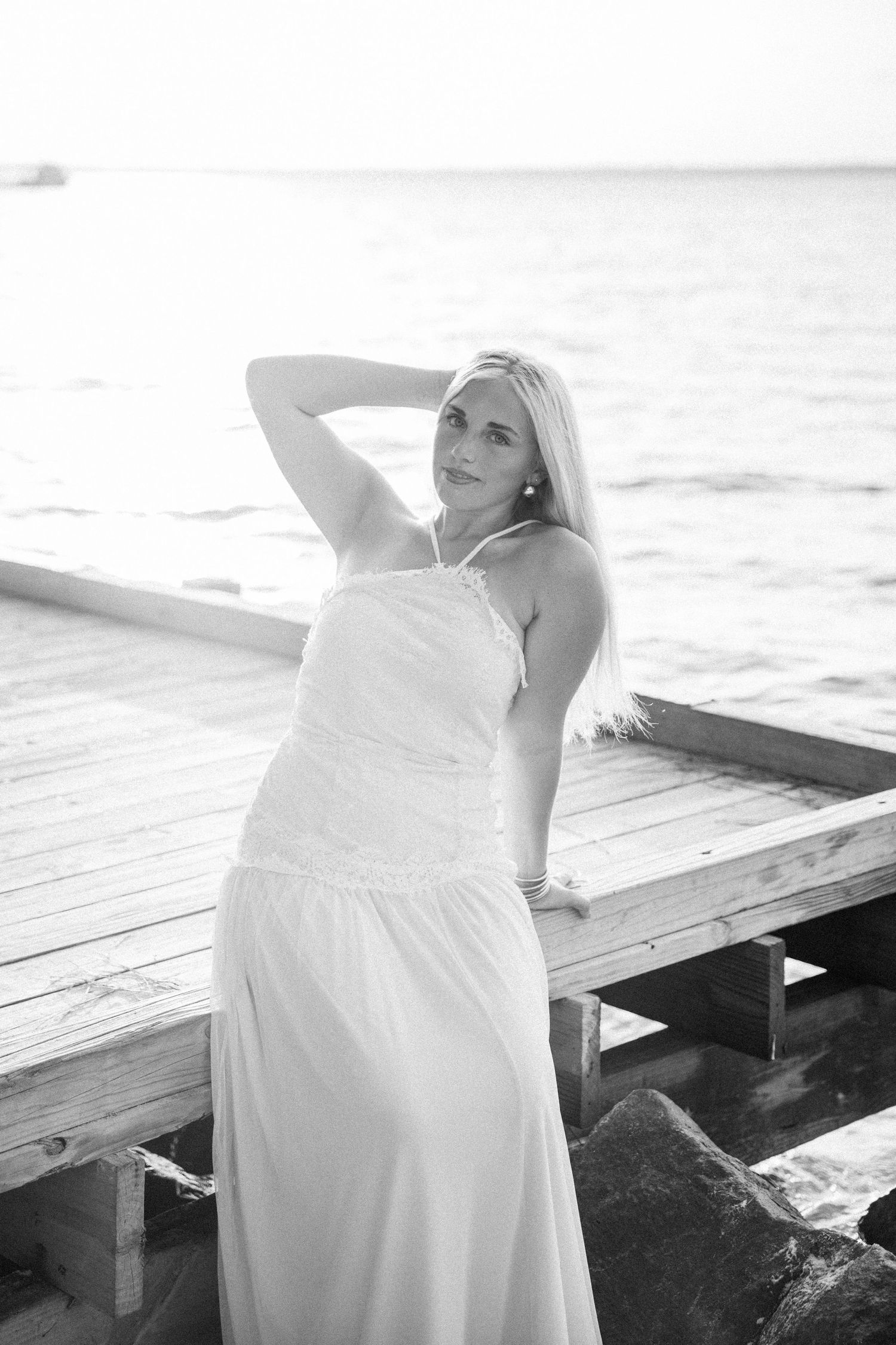 Black and white photo of a blonde woman in a white dress posing on a wooden pier by the ocean.