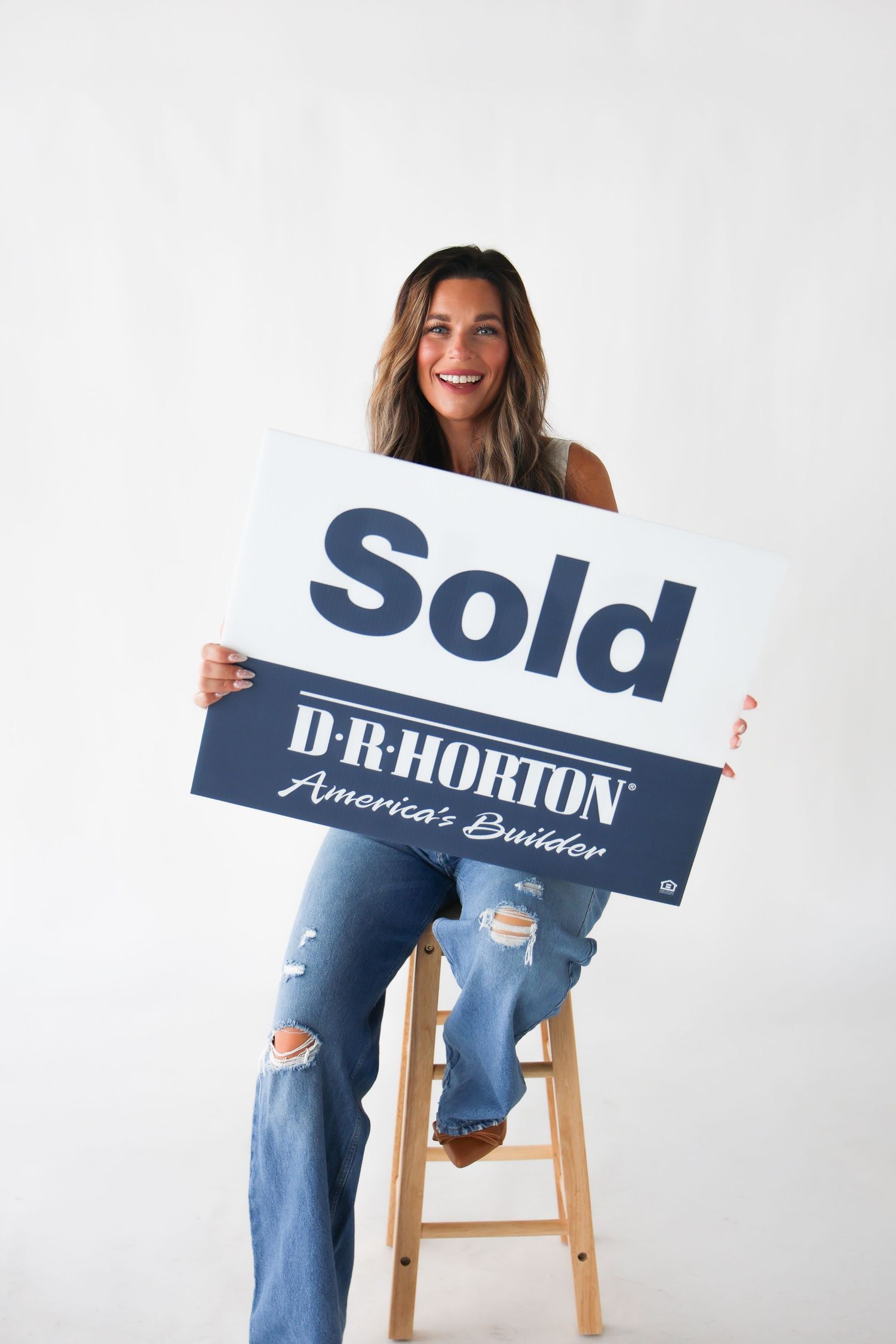 Smiling woman sitting on a wooden stool holding a D.R. Horton Sold sign, wearing ripped jeans.