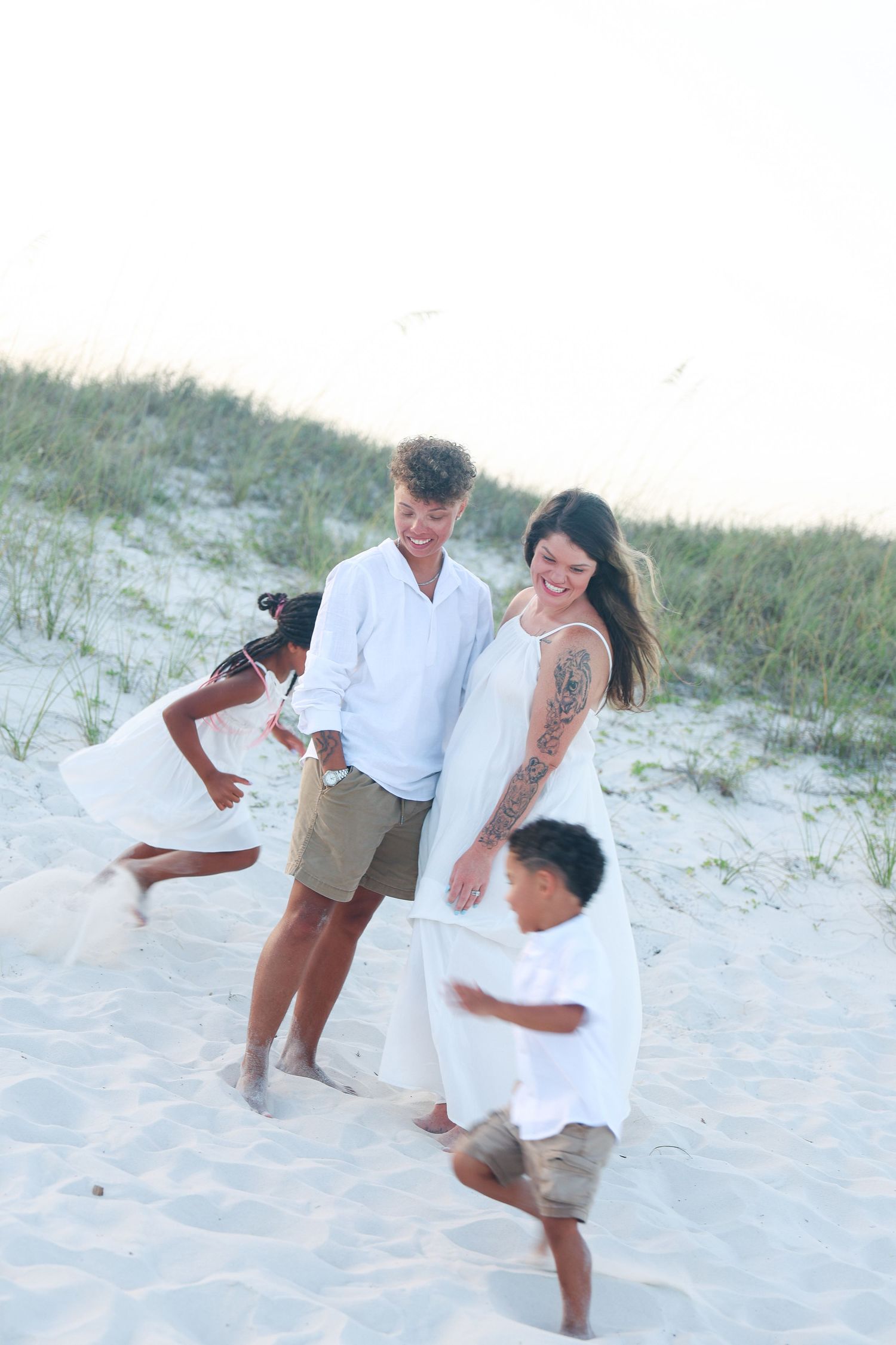Family of four in white outfits playing on a sandy beach, with two children running around their smiling parents.